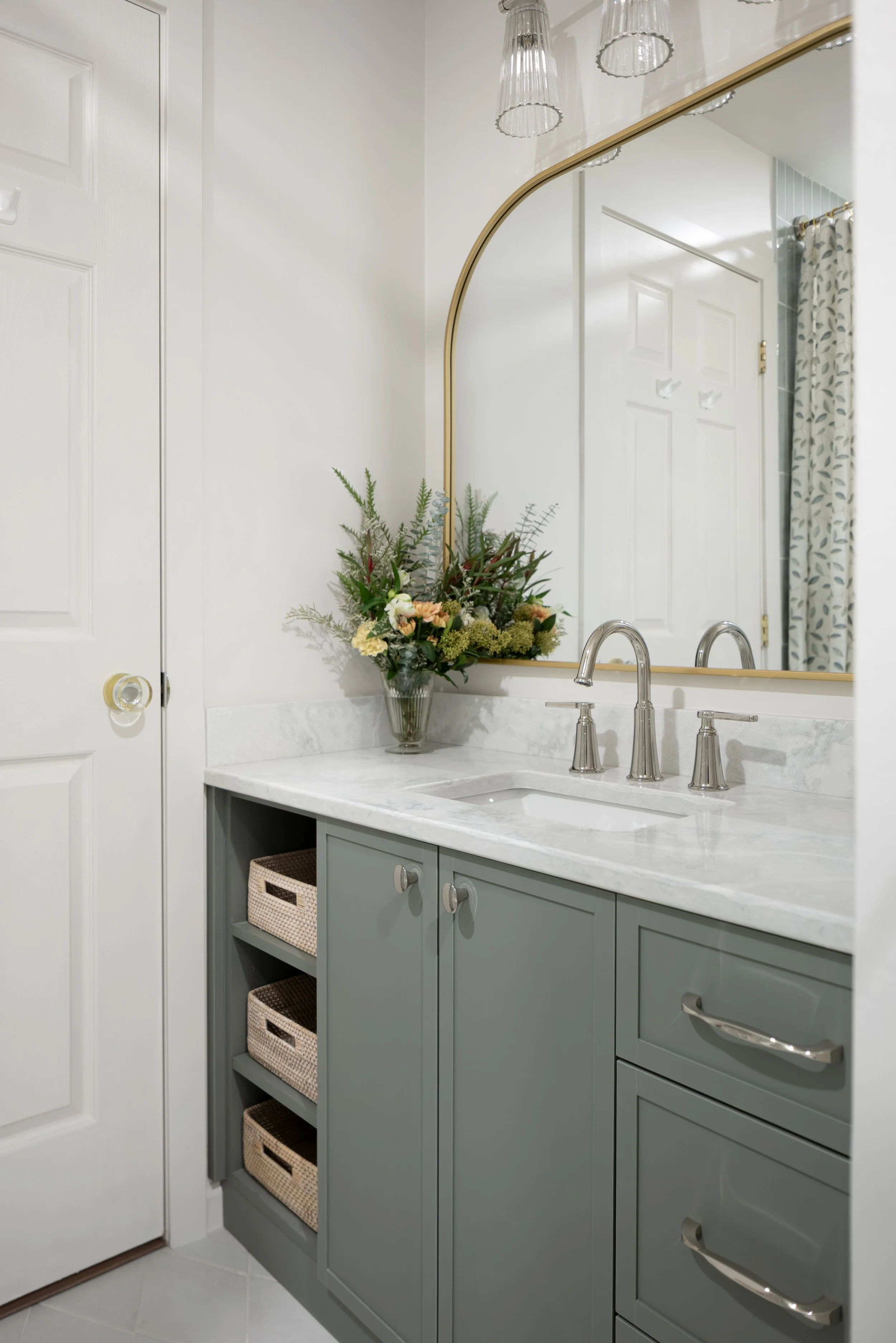 After Image of a bathroom reno that used a grey-green vanity. It also includes three woven baskets on the left bank of drawers. As well a beautiful, large vanity mirror with an arch on top and a thin gold frame which adds warmth to the all white room