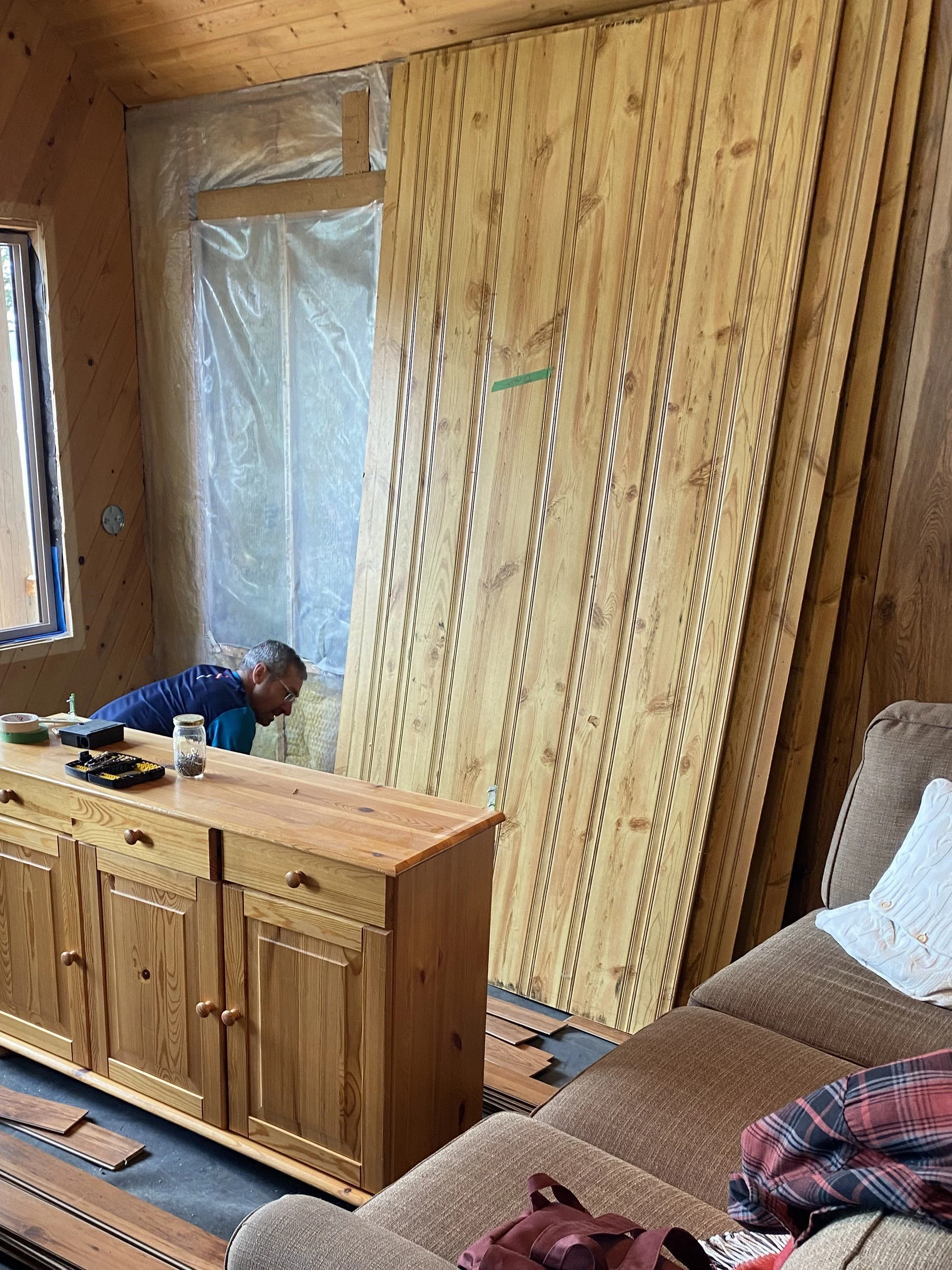 A wall that had a window on it has been filled in with insulation and plastic sheeting. Beside this a stack of about five tall 4x8' pine paneling sheets lean against the wall. They are ready to get installed on this wall as the wall has no material.