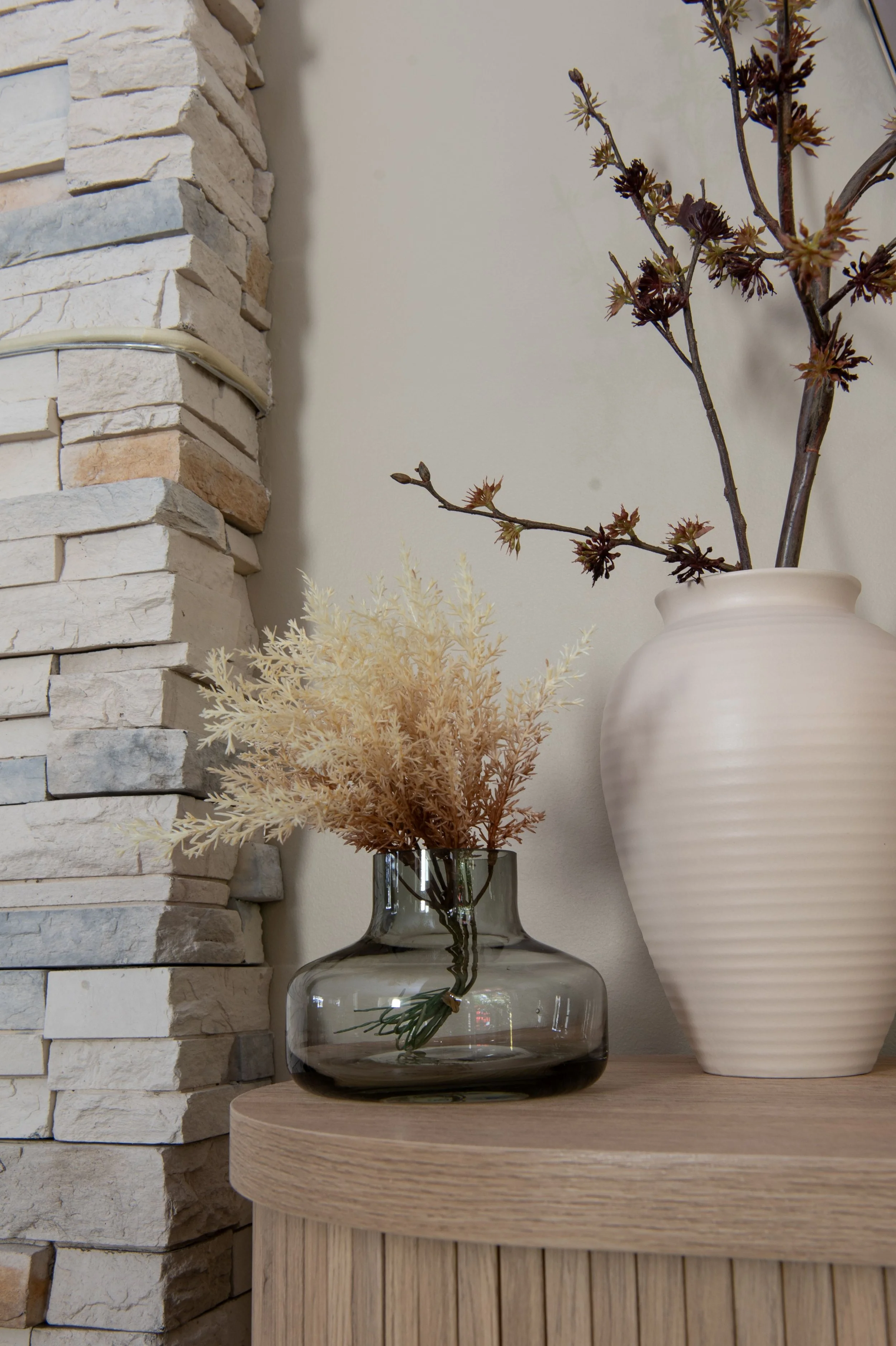 The edge of a neutral colored stacked stone fireplace is beside a low console table made of white oak and tambour wood. On top are two vases: one black glass from Torre and Tagus and one tall white cream oval vase from Renwil. Faux branch West Elm.