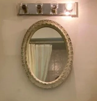 A none working vanity light (one bulb is on out of 4 bulbs) and the oval mirror is small with a traditional frame. The mirror is reflecting a white shower curtain, white shower rod, lowered ceiling and beige tile. It is a very plain looking bathroom.