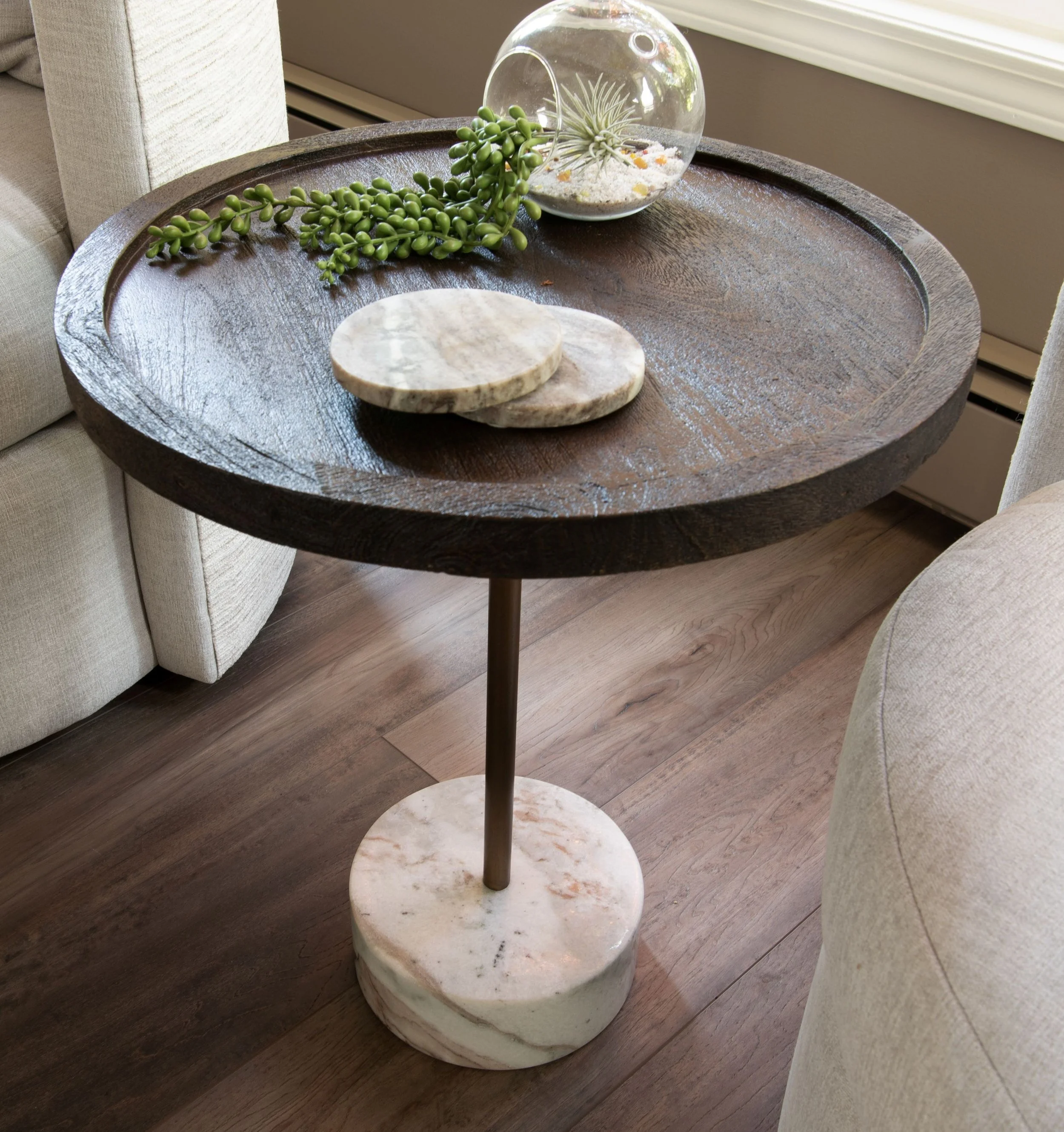 A short side table called Braiden by Renwil has an 18" brown stained wood top, gold post and marble base. It is a new side table in a living room next to new swivel chairs. The table has 2 marble coasters on top and a glass round vase with sand.