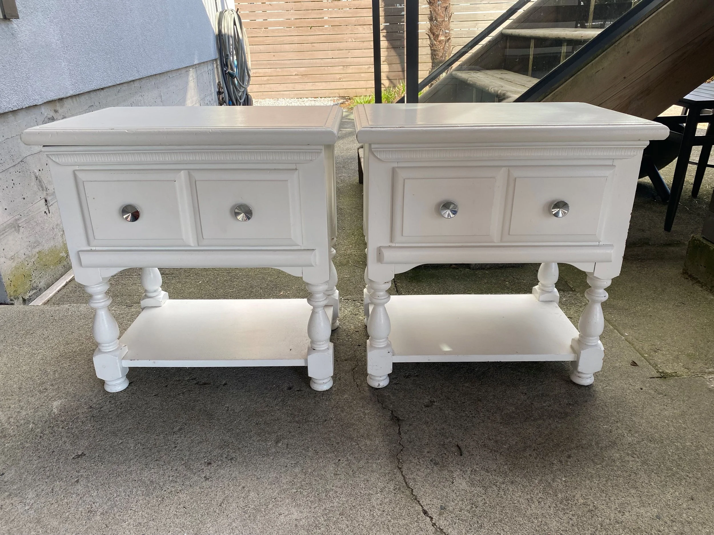 An identical pair of white bedside tables from the 1970s. Heavy and solid. Painted white top with one drawer and open to the base. Has round, curved turned wooden legs that are also white. Perfect for a DIY painting refinishing project.