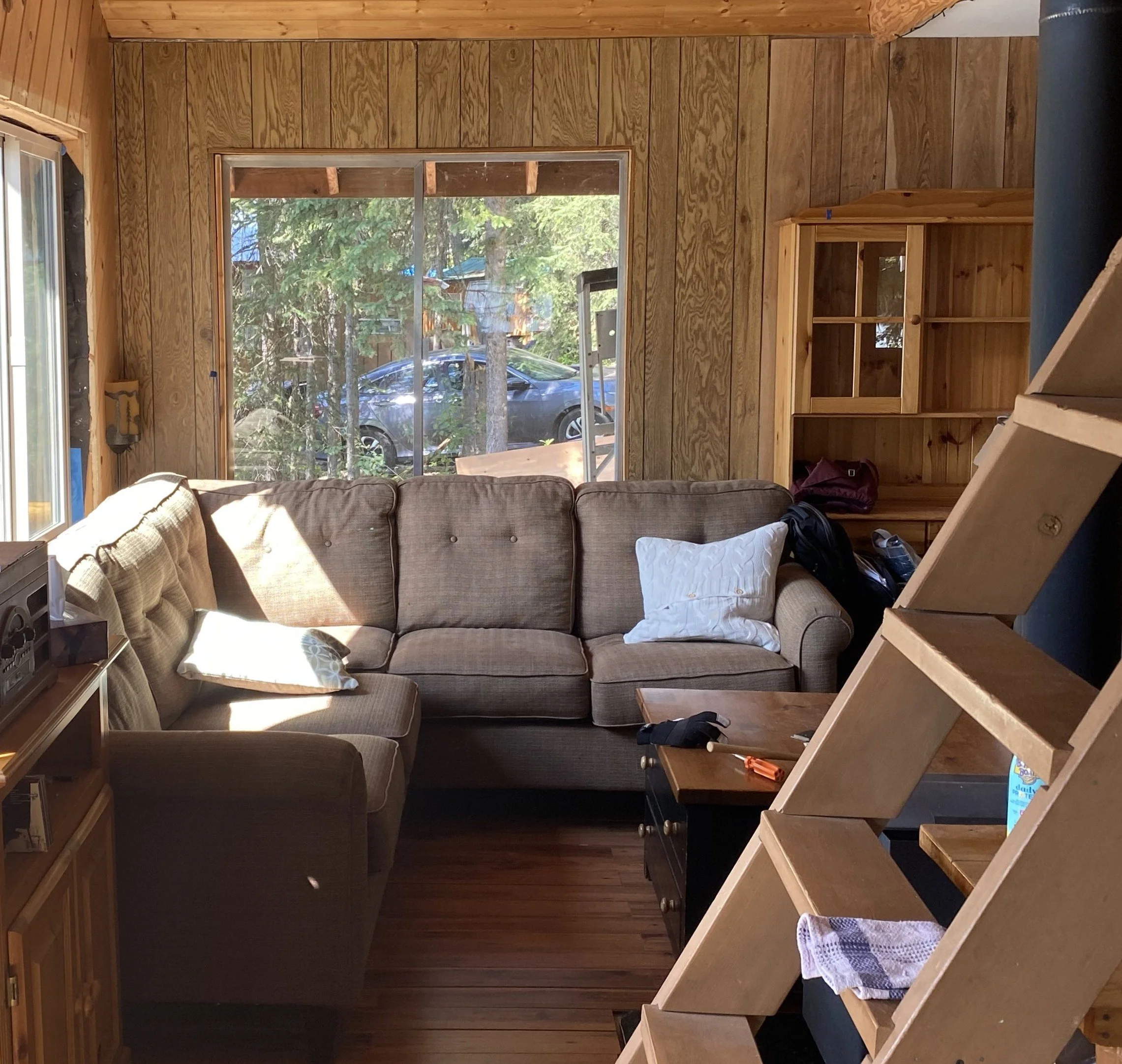 A large 48" wide window has been removed and the frame is open to outside. In the background are trees and a car in a driveway. In the cabin in front of the window is a large brown sectional. A ladder takes you up to the loft. Faux wood 4x8' paneling