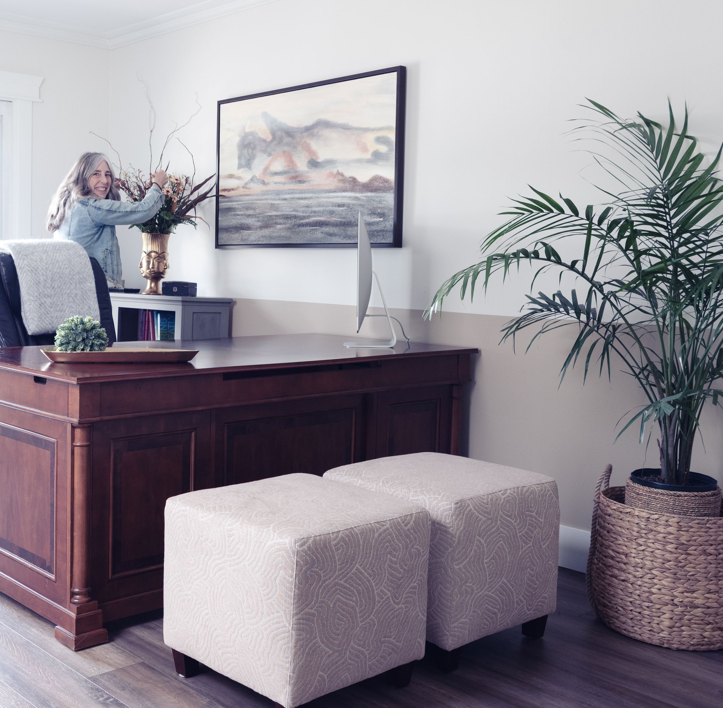An "After Photo" of a newly updated office space whereby the painted wall, new large artwork and ottomans and plant all help to make the space feel intentional. The designer, Kelly Grimes, is rearranging the flowers in the vase in the background.