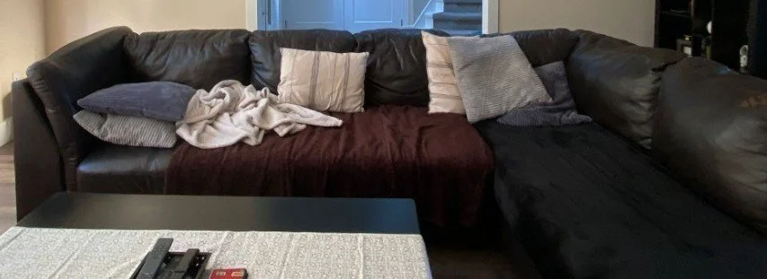 Before photo of a brown leather sectional which has dog blankets on it for protection and super flat sofa toss cushions doubled up for comfort when laying. Also a dark brown living room table is in front of the sofa.