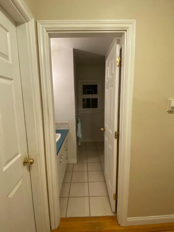 A late 1950s dated bungalow bathroom entry. The door is open and there is very little light coming in. The floor are square 12x12 tiles and the vanity counter is a bold teal green color. Everything else is off-white and not inviting.