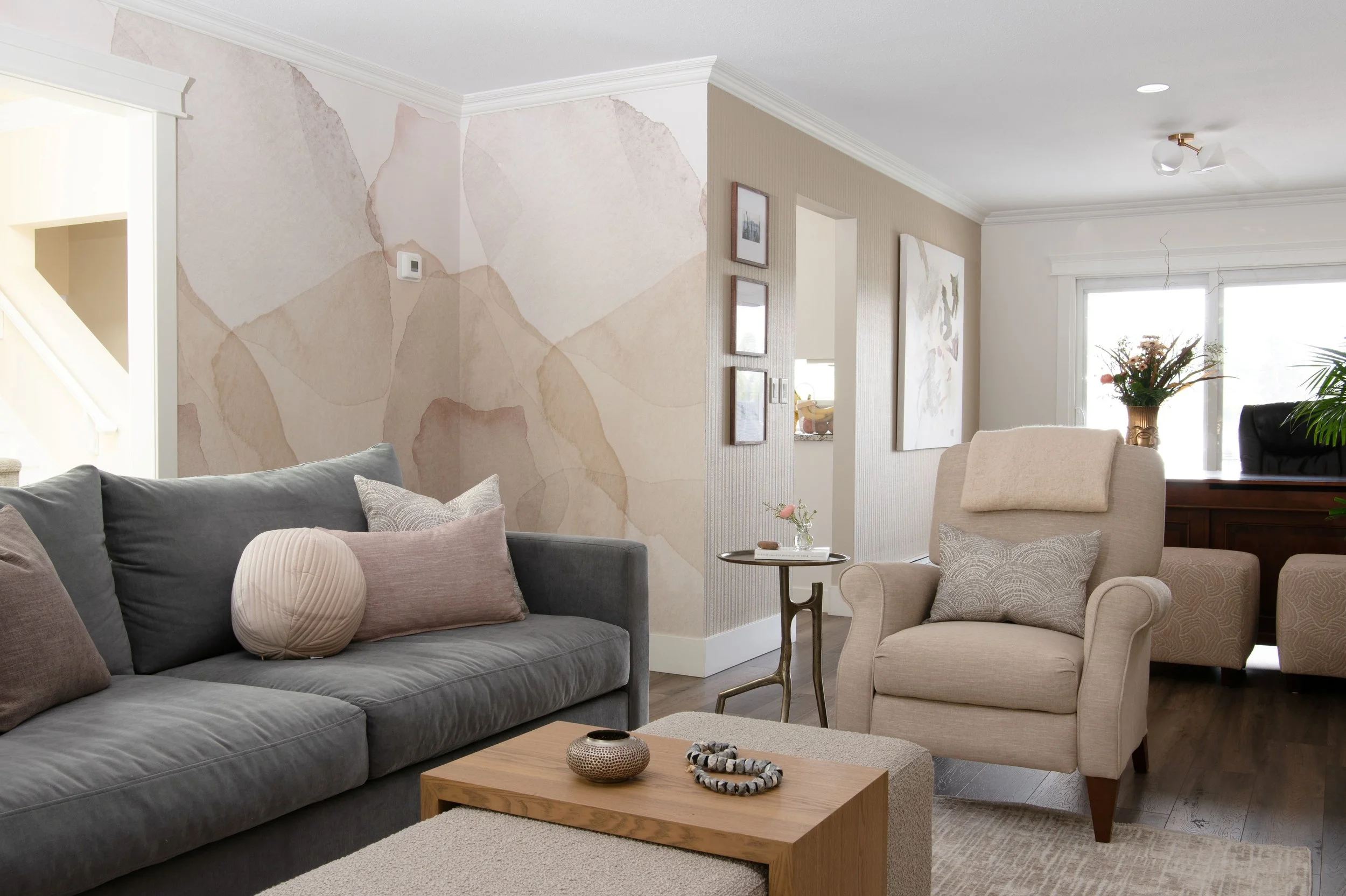 An updated living room has a wall mural of large organic flowing abstract petals in soft shades of pink and sand color. In front is a dark blue-grey sofa with pinky toss cushions and also a reclining chair updated in a neutral beige fabric.