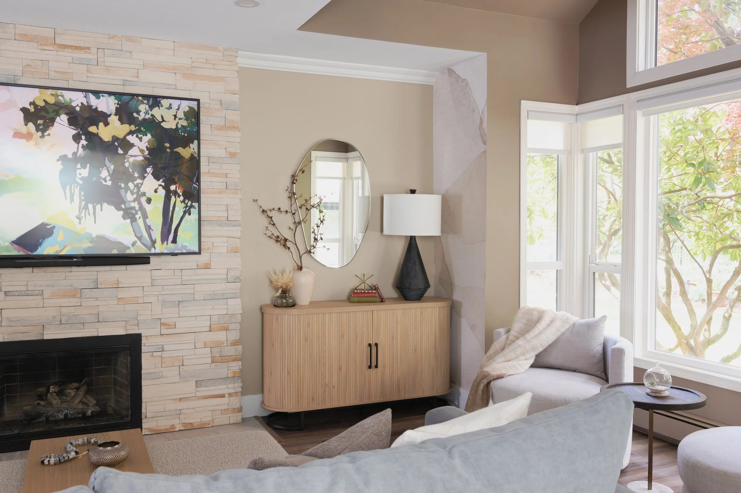 A newly updated living room features a stone stacked fireplace in cream, grey and orange colors. Beside is a tamboured, white oak side table from Wayfair. Black lamp and accessories sit on top. An organic shaped mirror is on the wall behind.