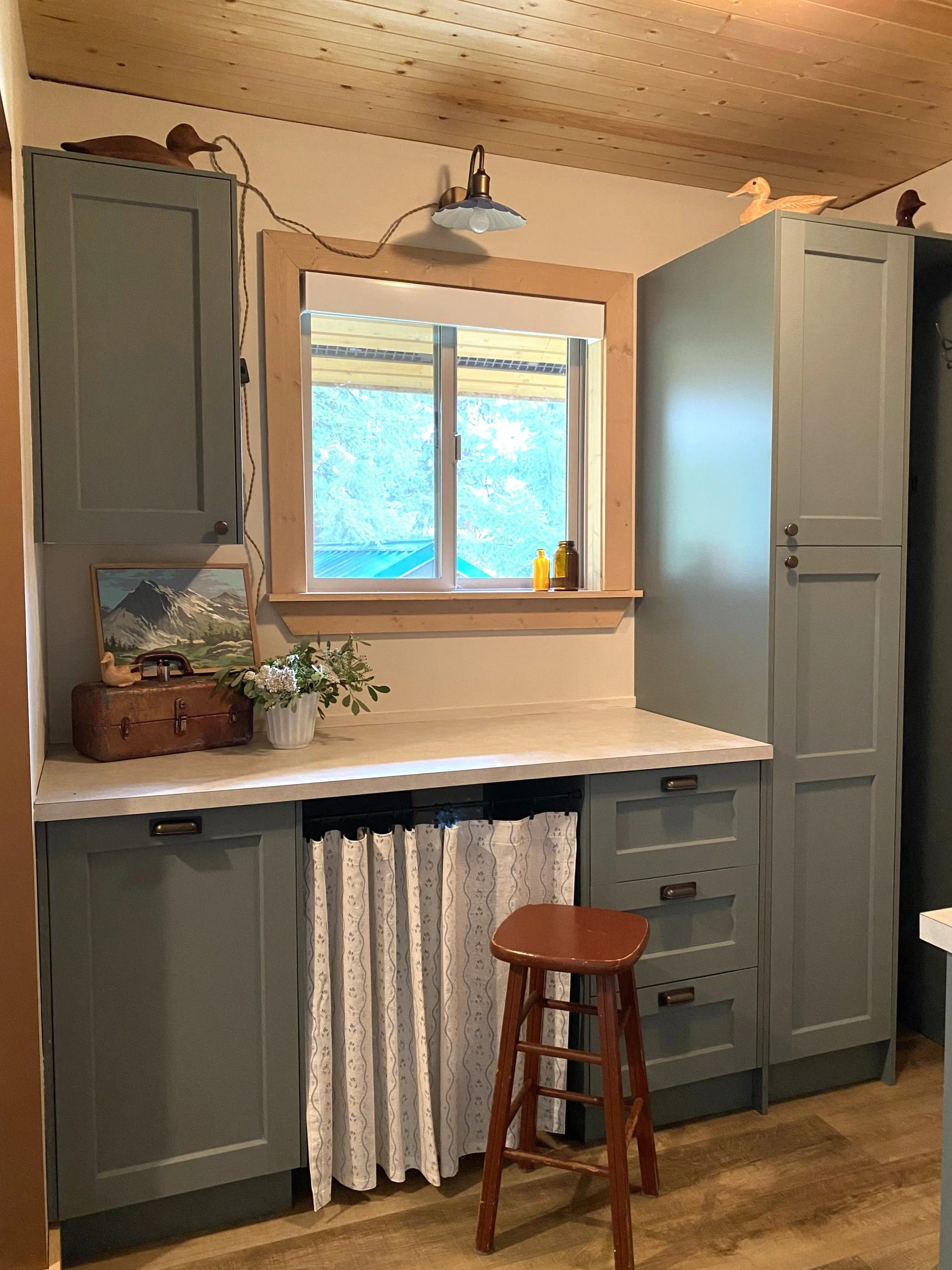 A completed renovation of a storage room with a cottage feel. Green IKEA cabinetry, tall pantry, Lee Valley vintage bronze hardware, light over the window, duck collection on top of cabinets, rustic red stool, white linen curtain hangs below counter.