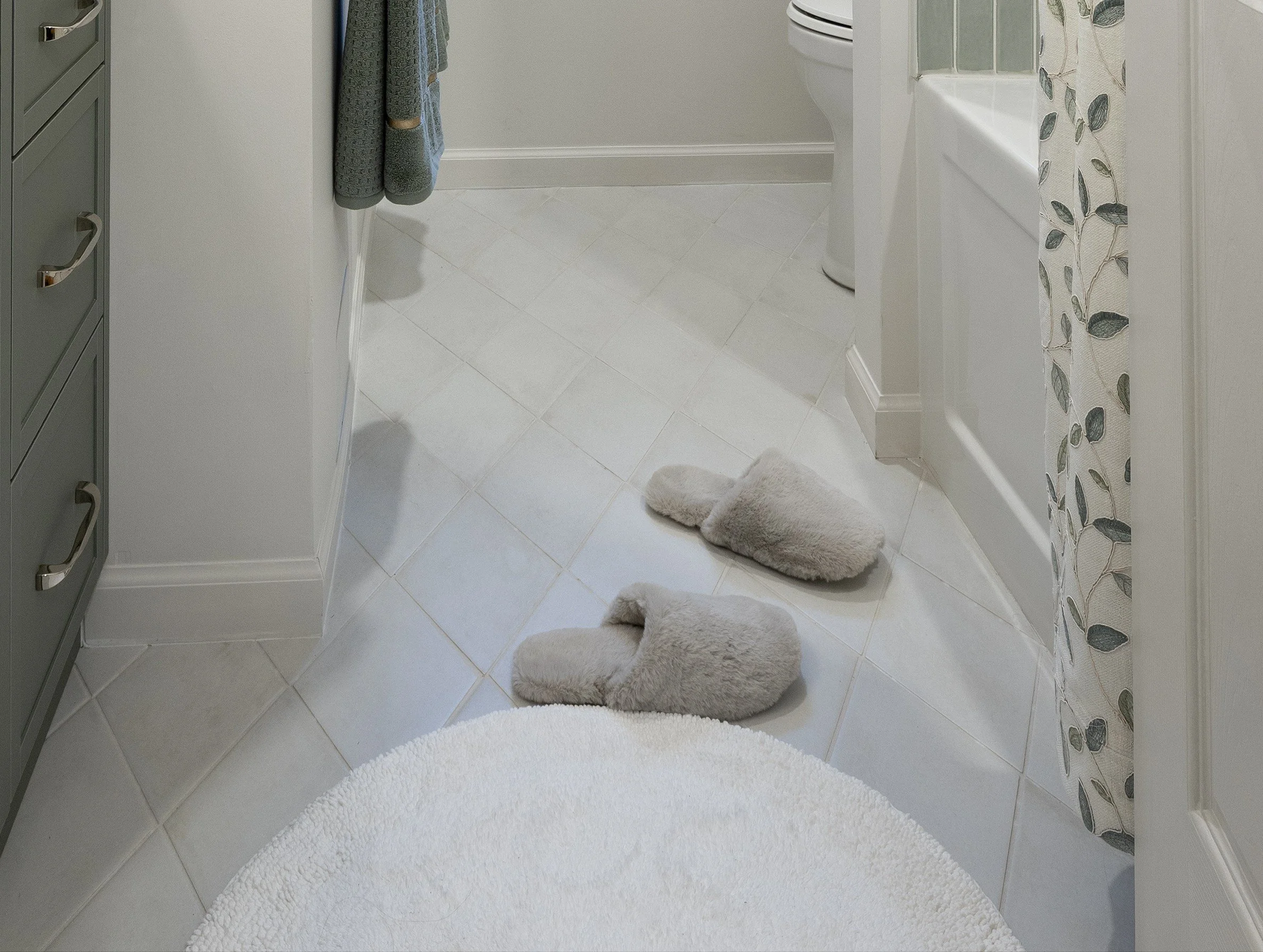 A bathroom floor features a white diamond tile floor (Ossidi tiles from Olympia Tile) along with a cozy round white bath math and fuzzy spa slippers.