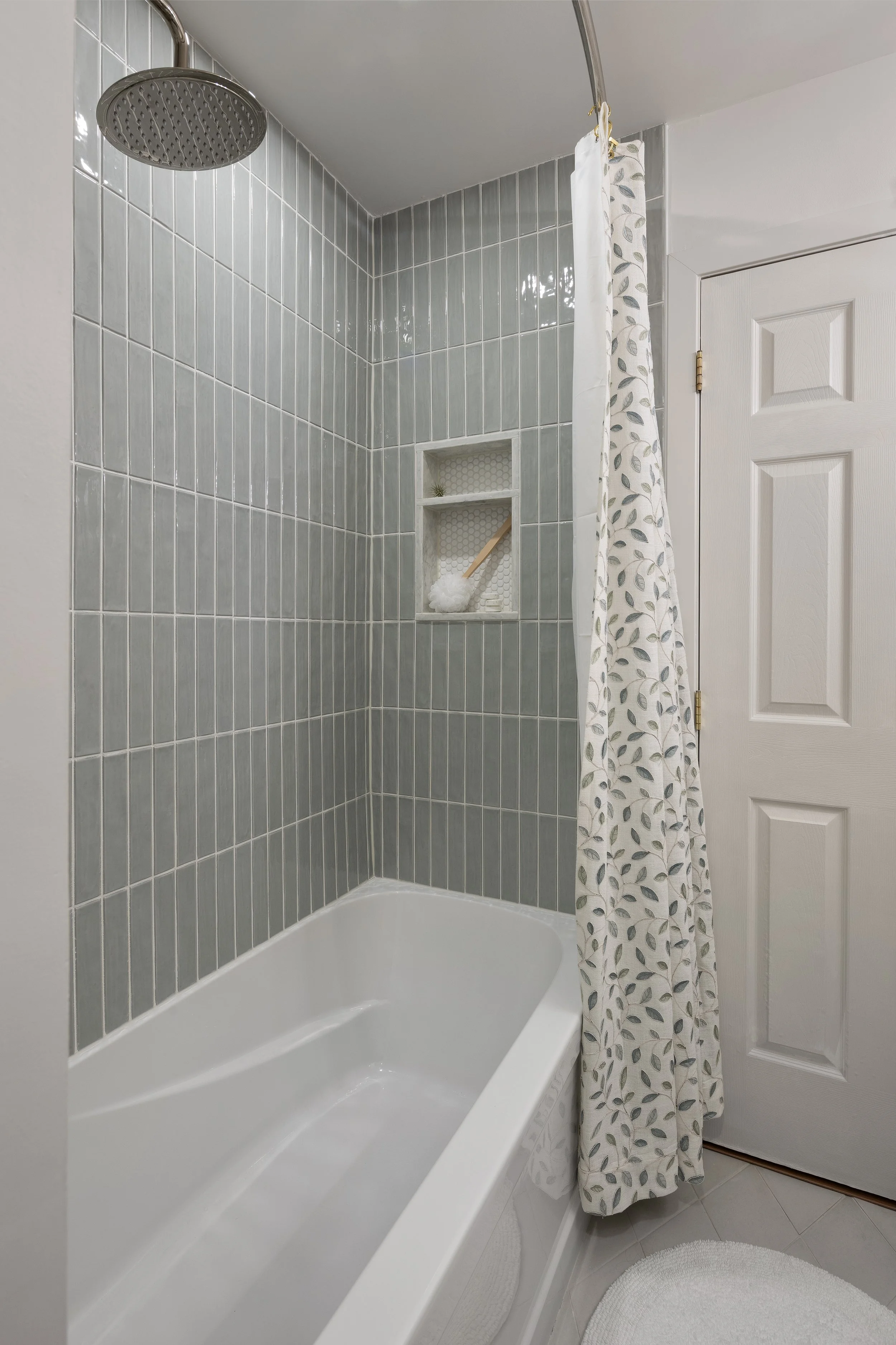 After image of a newly renovated bathtub and shower. Soft green spa-like colored rectangular tiles are in a modern stacked pattern. The shower niche is small at the end and is framed in quartz with a quartz shelf and white penny tile on the back.