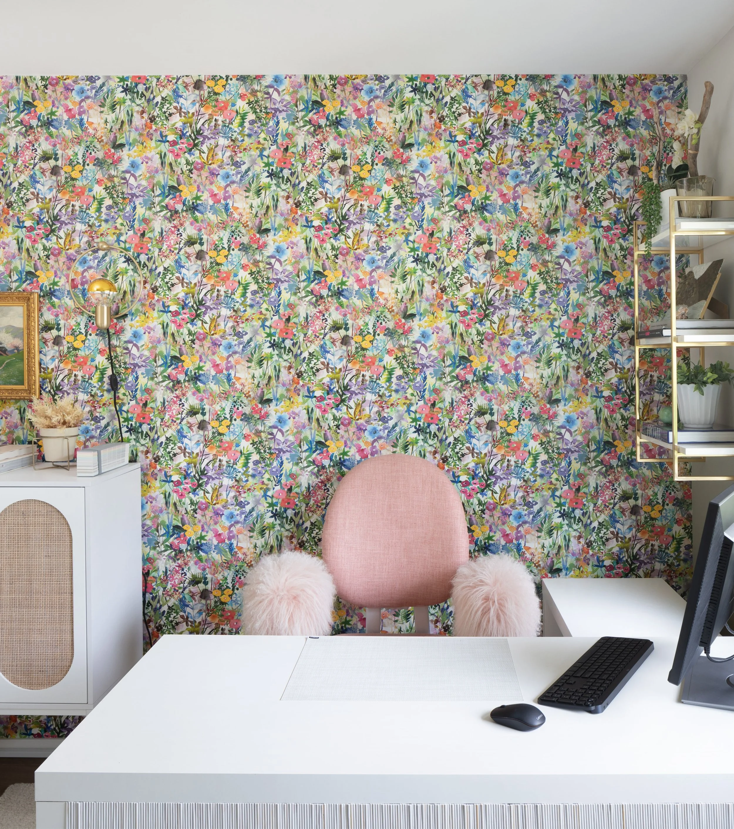 A home office space with vibrant flower wall paper. Also vintage gold picture frame and gold wall sconce are on the wall above a white tall storage cabinet. Behind the white desk sits a light pink office chair with fluffy light pink mongolian fur arm