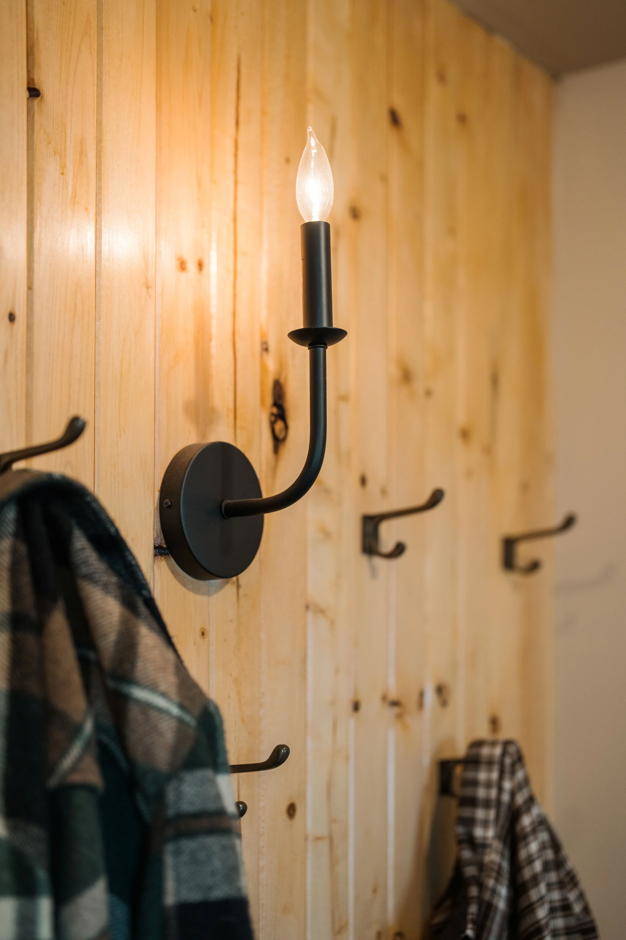 A single black candle wall sconce with only a light bulb at the top (no shade) illuminates the pine wall and vintage brass coat hooks. Two plaid jackets hang separately on the hooks. One is a vintage green plaid coat and the other is brown plaid coat