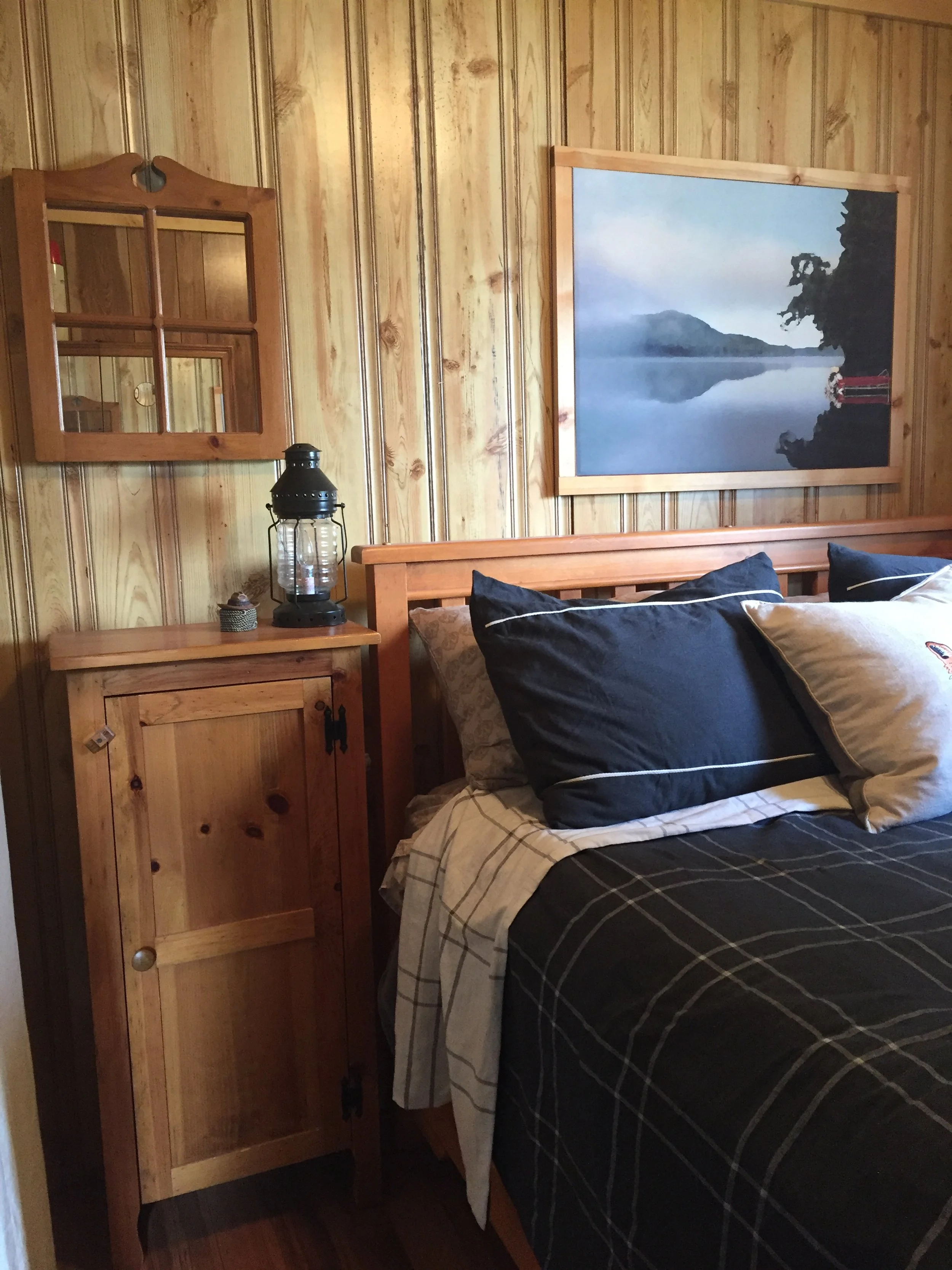 A pine paneling wall in a cabin is behind the queen headboard. To the left of the bed is a narrow pine dresser with an electrical vintage lantern on top and a pine heart mirror with four mirror panes sits on the wall above the dresser.