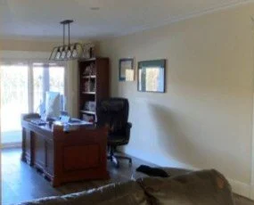 A "Before" photo of an office, which is in an old dining room, so the original dining chandelier (an open 4 light linear in a black frame) sits above the desk. The large dark brown desk chair and the tall brown bookcase in the back all look dated.