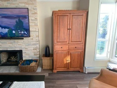 Fireplace before photo shows a large dated and rectangular orange-brown wardrobe furnishing. Squeezed in a space beside the fireplace, it looks like it doesn't belong there. Woven baskets and boxes sit nearby.