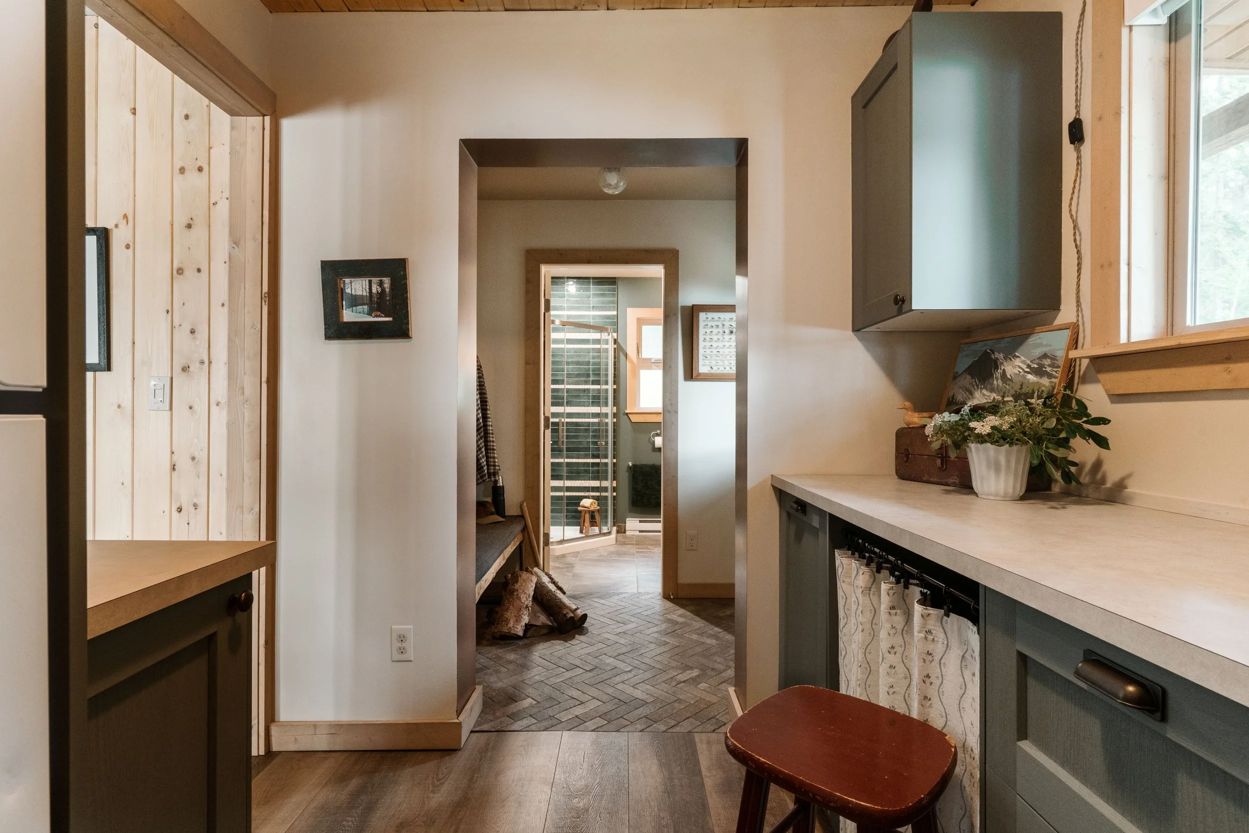 View from inside a storage room looks straight ahead to a doorway opening of the entry room adjacent. And beyond that, continuing is a doorway (with it's door open) leading into a bathroom that has a green and white horizontal striped shower.