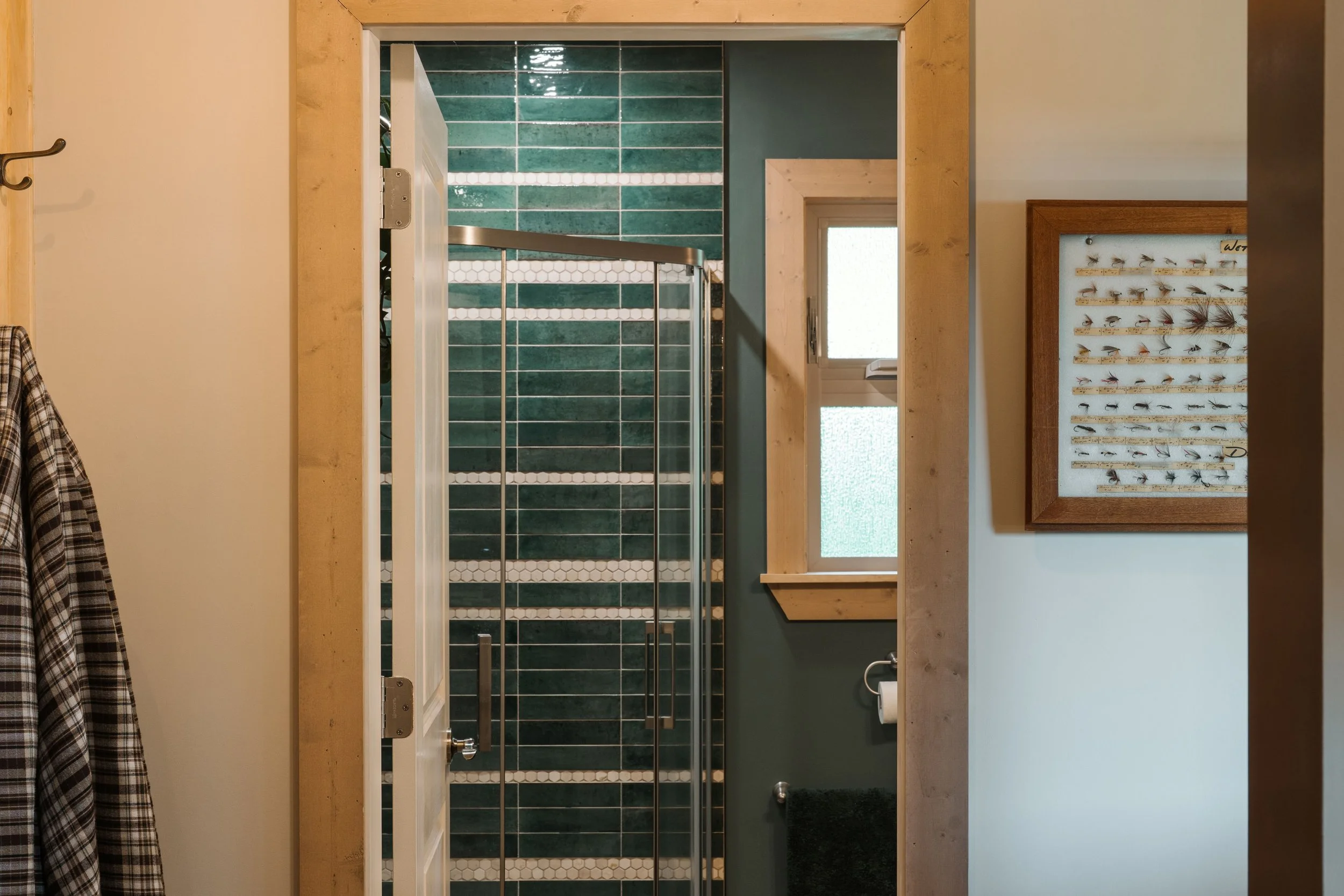An entry into a cabin bathroom has a coat hook to the left of the entry and a vintage fly fishing ties collection framed and hung on the wall to the right of the doorway. In the bathroom is a green and white striped tiled shower with a window trimmed