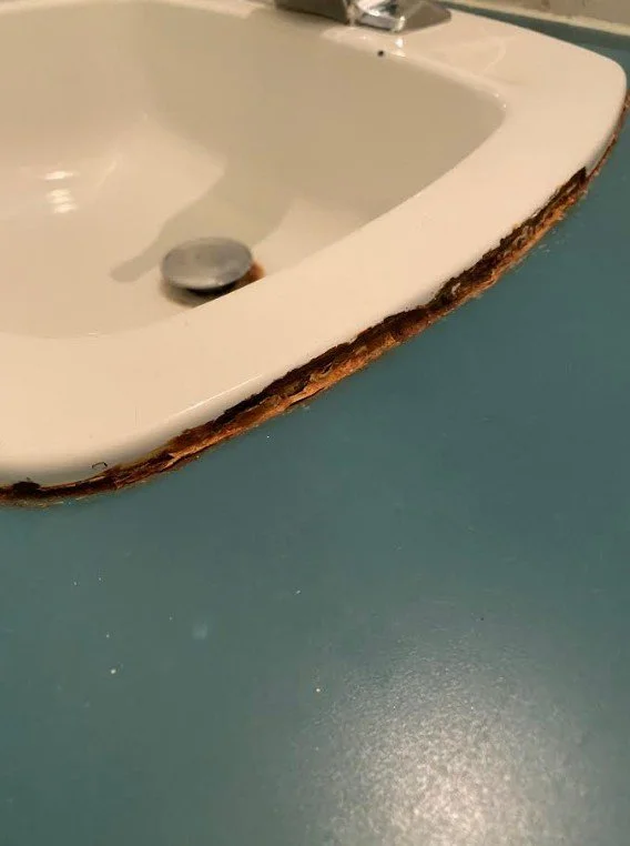 An original bathroom sink is outlined with rust where it meets the old laminate counter top