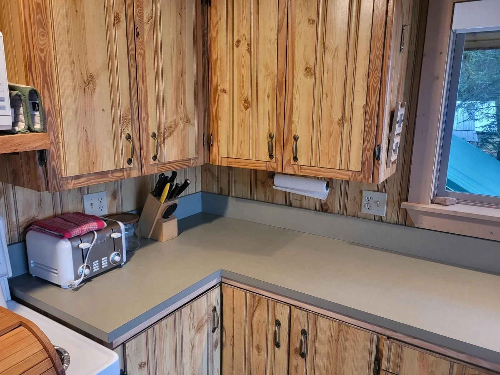 Sage green laminate countertop in a rustic cabin with pine cabinets and vintage brass handles for hardware. A knife block and toaster sit on the counter. A paper towel rack hangs from the upper cabinet.
