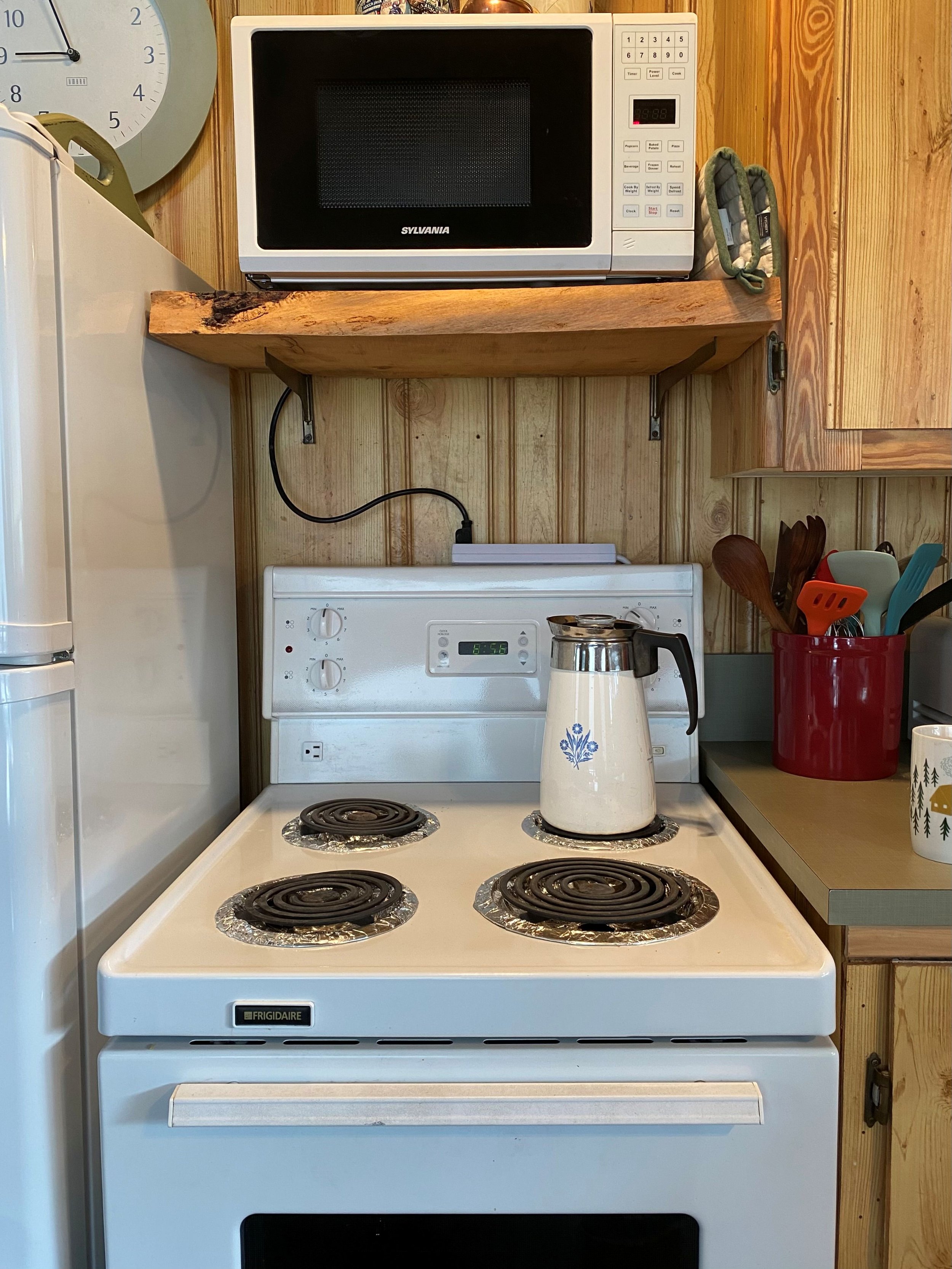 A 24" white stove in a cabin has a make-shift live edge shelf above it holding a white microwave. The microwave cable hangs down and is plugged into a cable bar that sits on the stove.