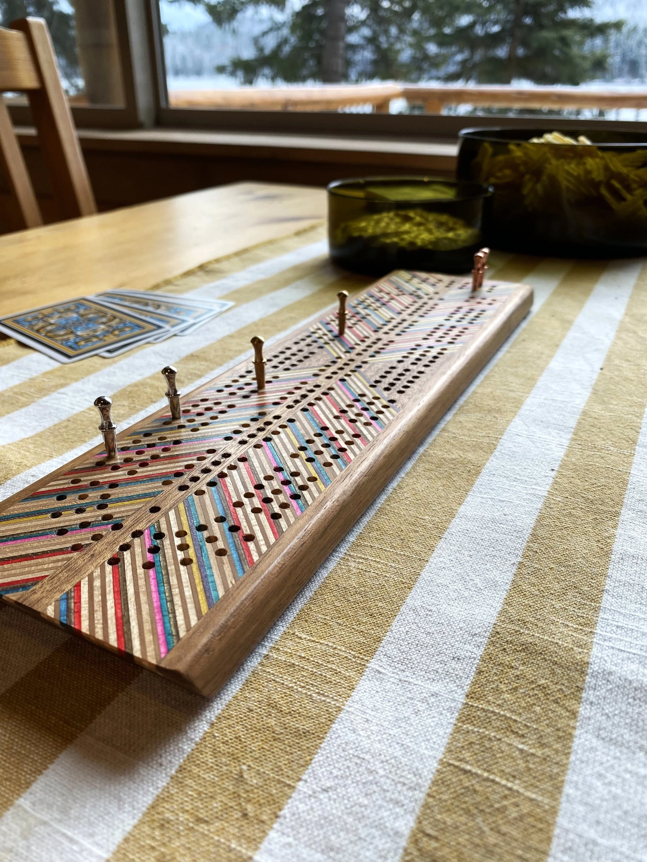 A very cool handcrafted crib board. It is made using walnut wood and skateboard bits that make it colorful on top, with an angled chevron design. Silver and gold pegs are in the holes. A vintage green smoked glass chips and dip bowls are shown.