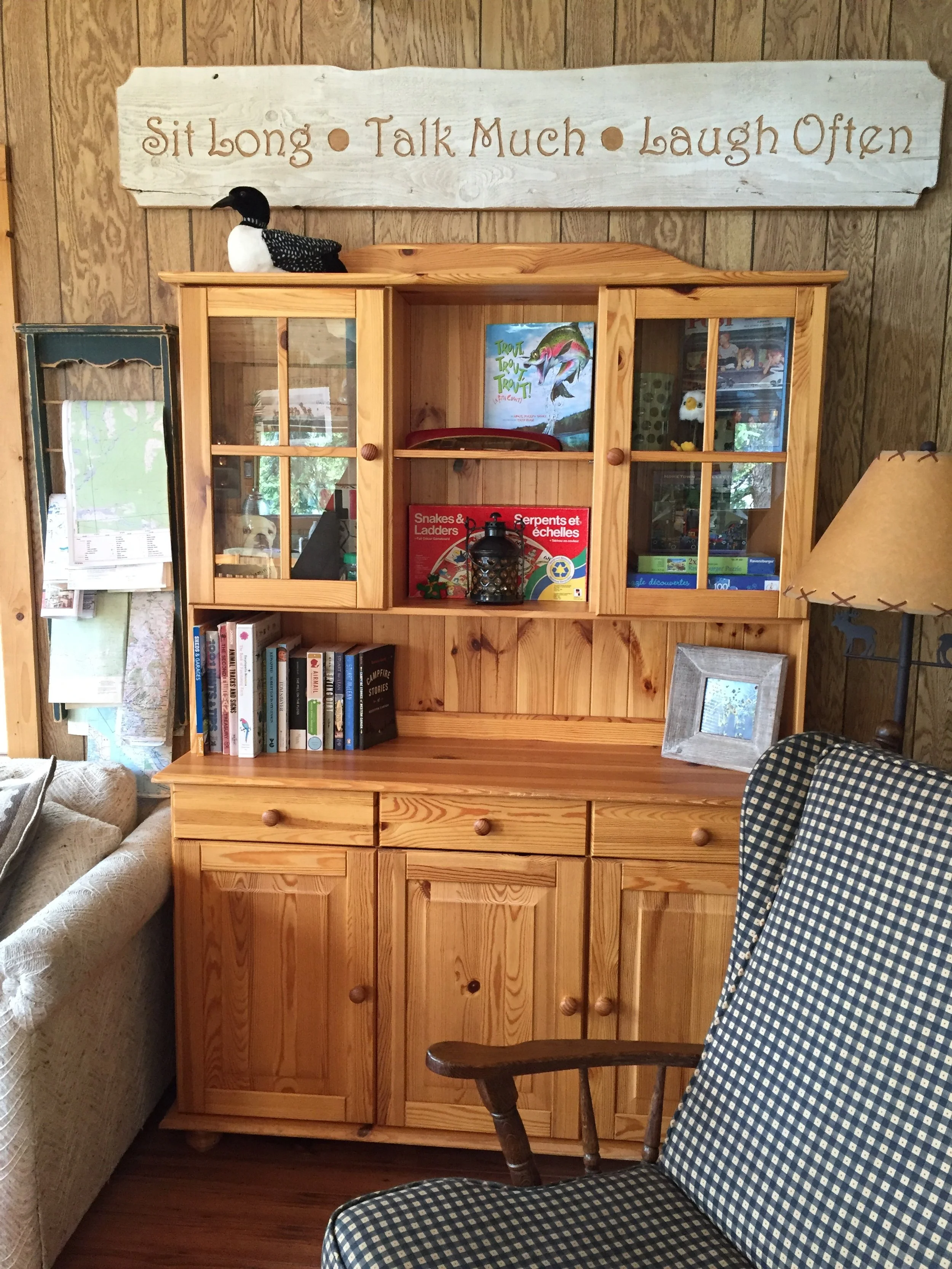 An old IKEA pine wood sideboard and hutch in a cabin setting. A hand made wall art sign sits atop and it reads "Sit Long - Talk Much - Laugh Often"