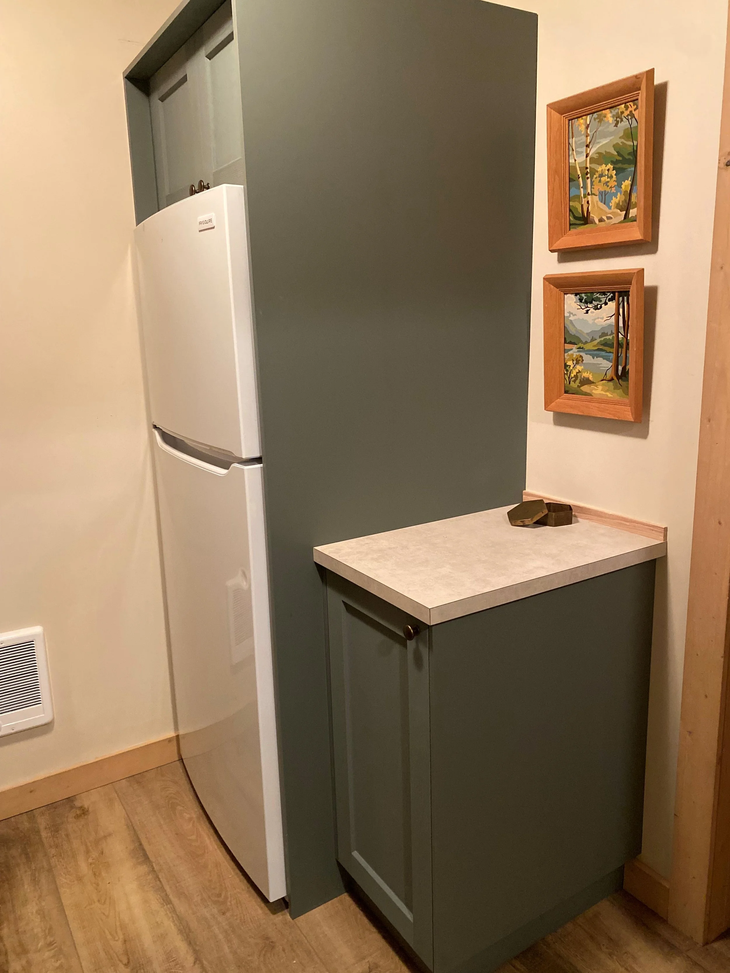 White fridge is inserted in a green fridge cabinet with upper cabinetry above for more storage. A wall mounted heater is nearby to keep the space warm. Vintage wood framed paint by numbers wall art is hung and brings a sense of nostalgia.