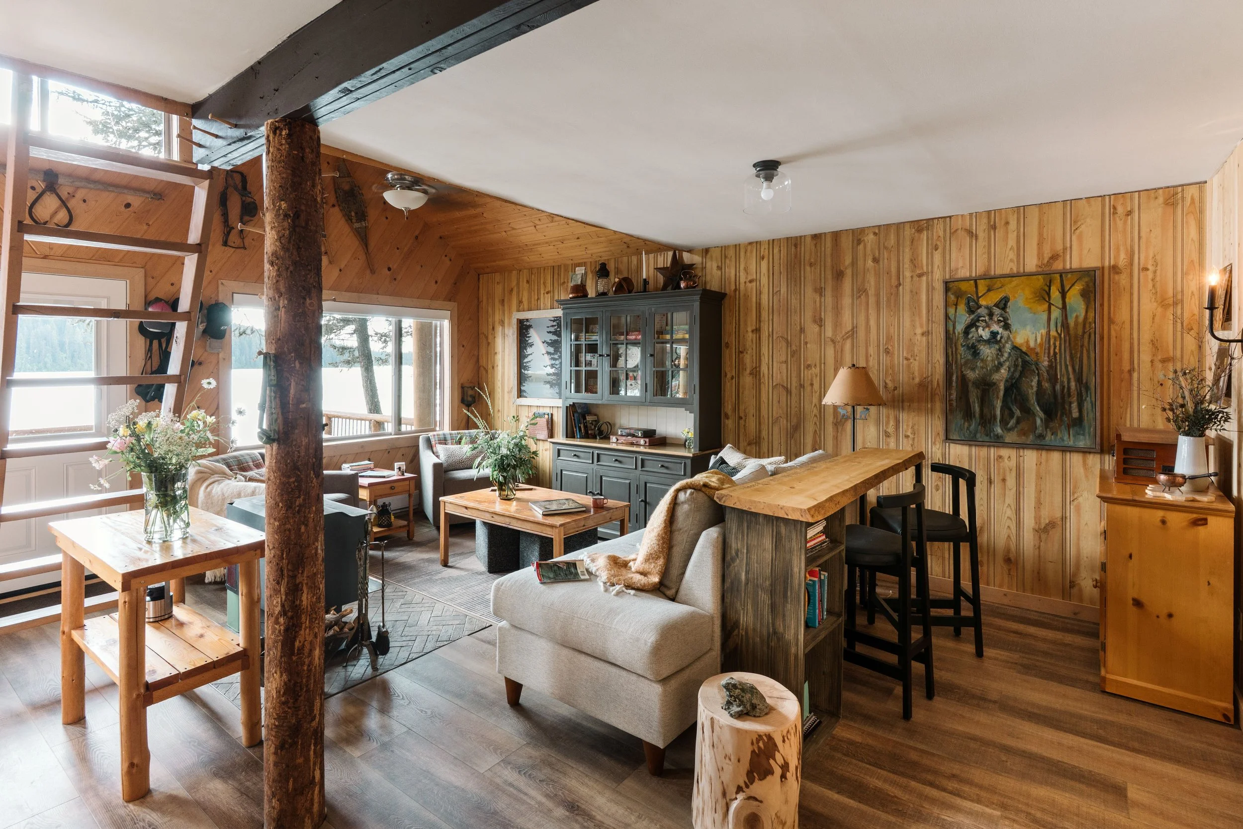 A cabin living room looks well appointed with new furniture and rustic pine accents. A wolf painting suits the room. An old tree acts as a post taking the load. The walls are angled pine tongue and groove and another wall has faux pine paneling 4x8'