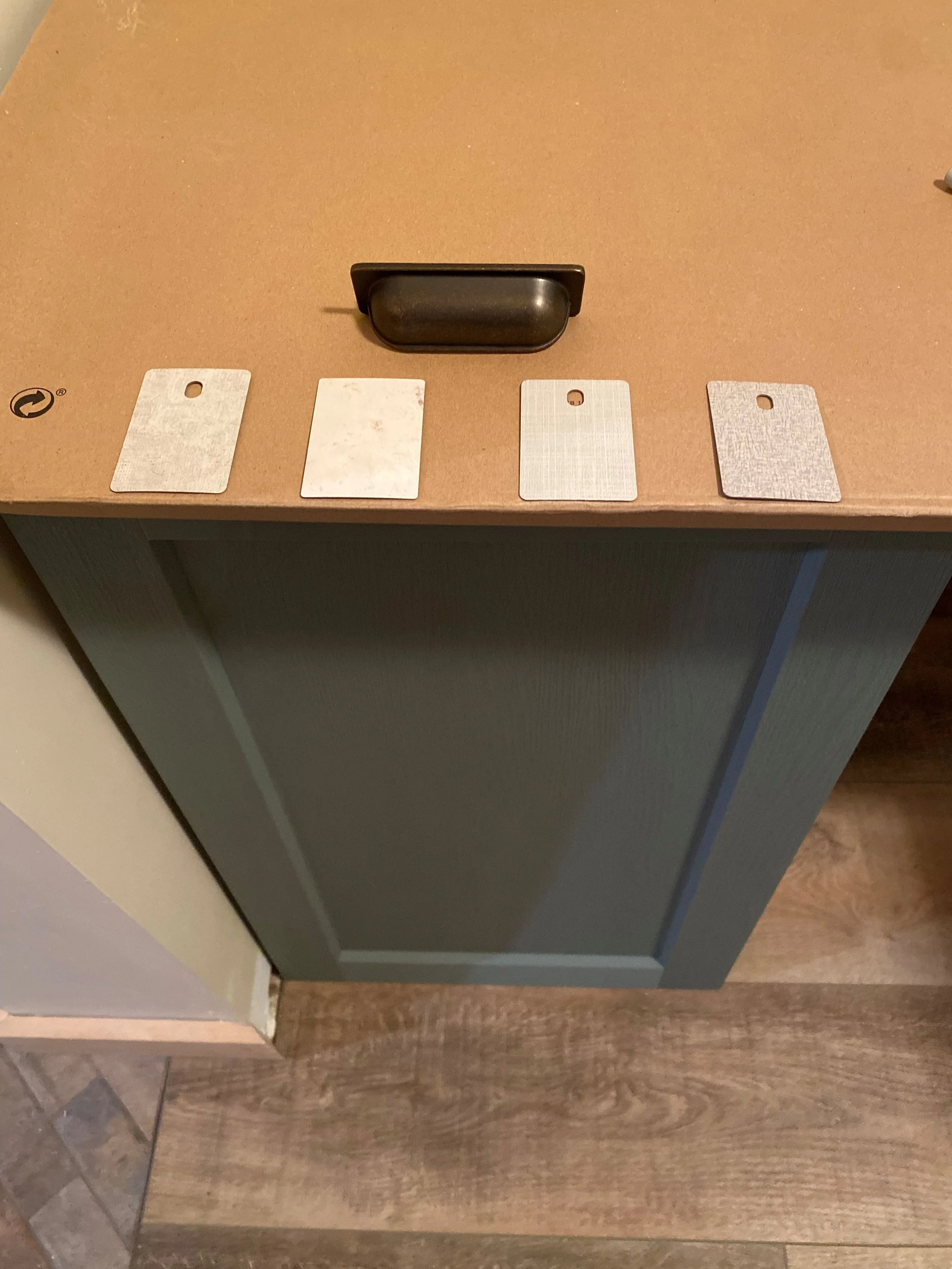 Four laminate counter samples are laid out above a cabinet to determine which one will work best with the vintage brass hardware, white/grey walls, green cabinetry and rustic Fuzion LVP flooring.
