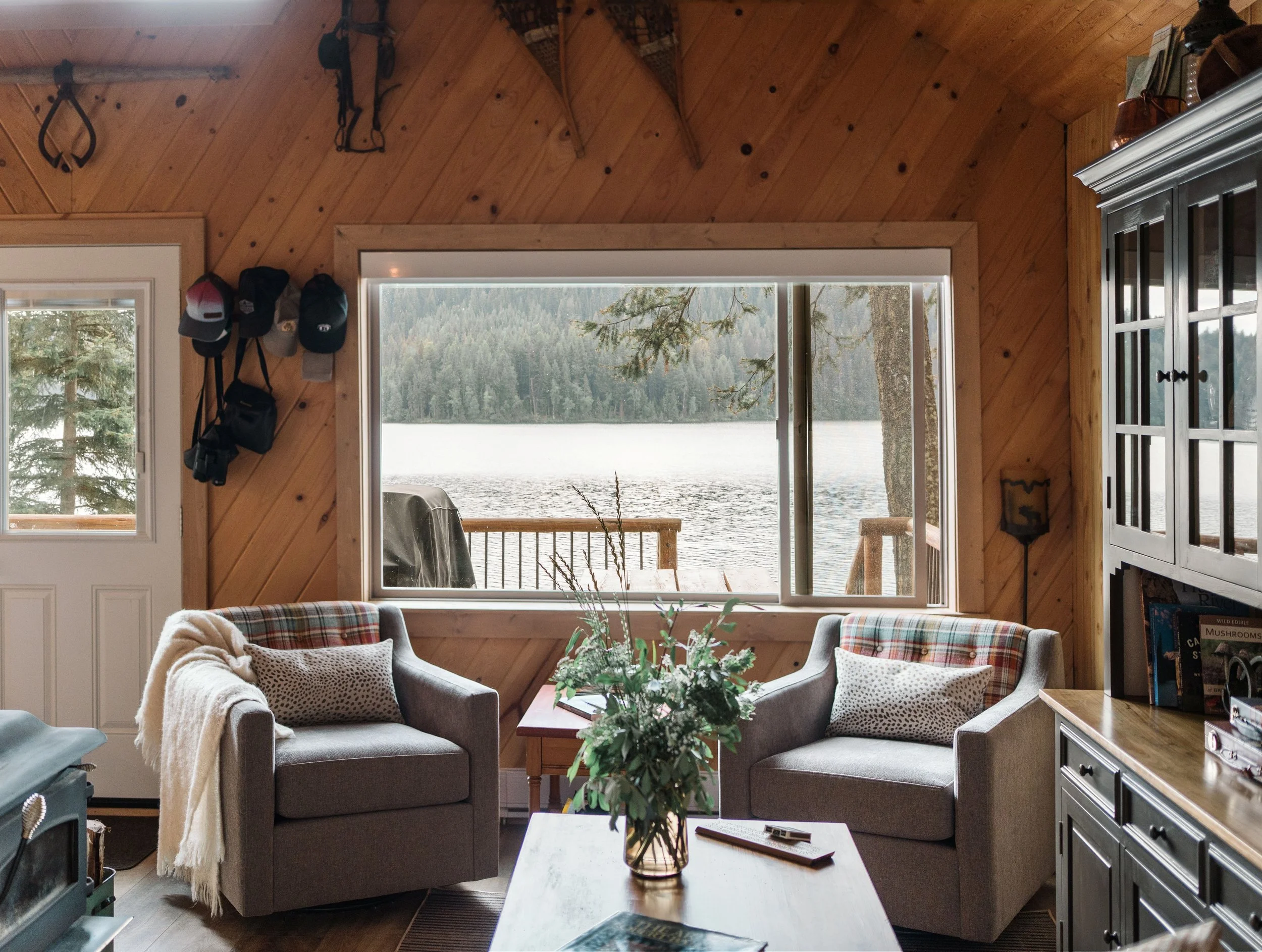 Two swivel chairs sit centered in front of a large window that has a view out to a lake and a mountain across from it. The setting is within a pine rustic cabin that has been updated with modern furnishings. Antiques rest on the wall as decor.