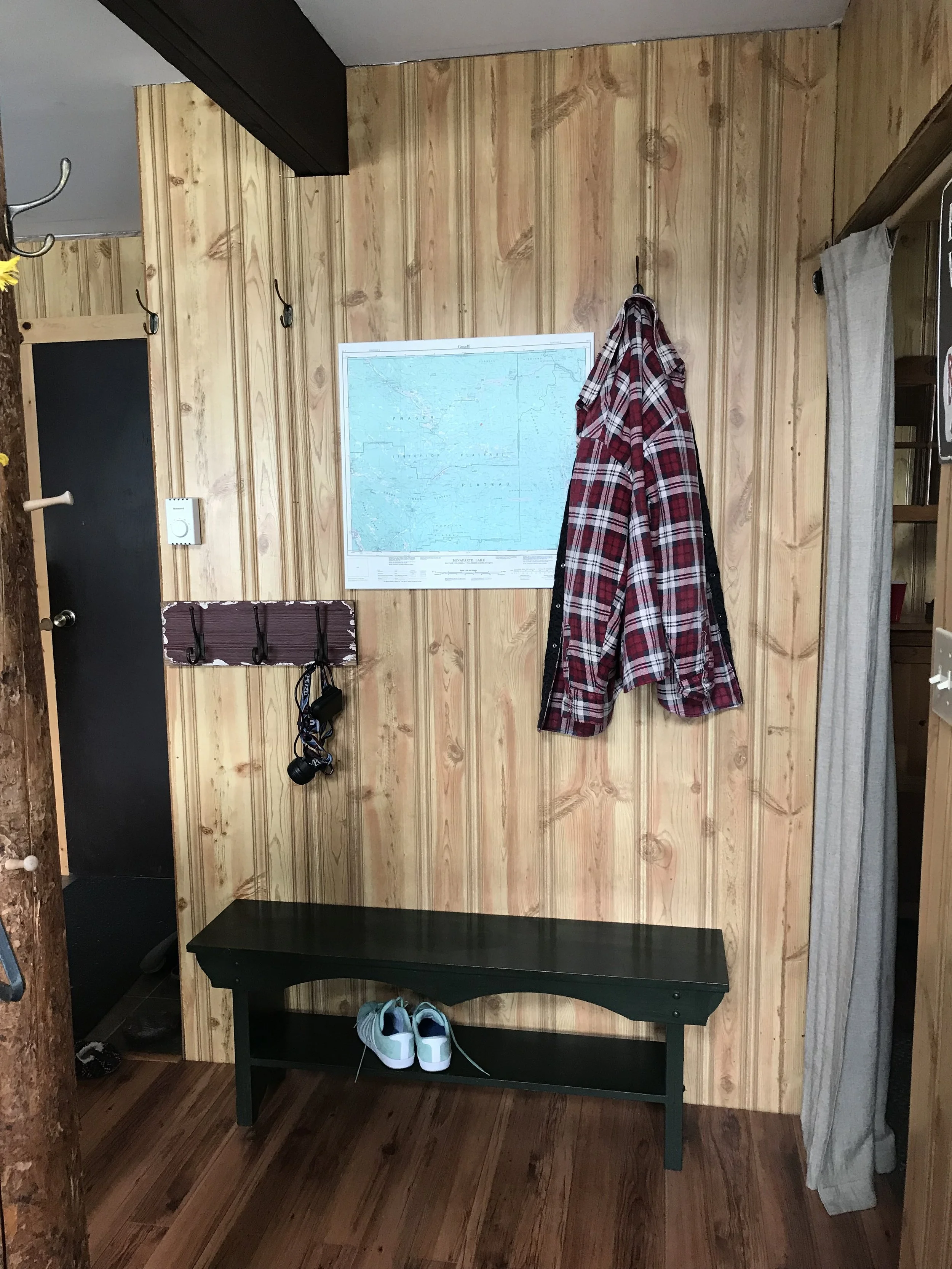 A wall with faux pine paneling juts out. Behind it is a closet space (can't see it) and an entry door. To the right of the wall is a doorway with a curtain door leading into a bedroom. A map and plaid coat hang on the wall. A green country bench sits