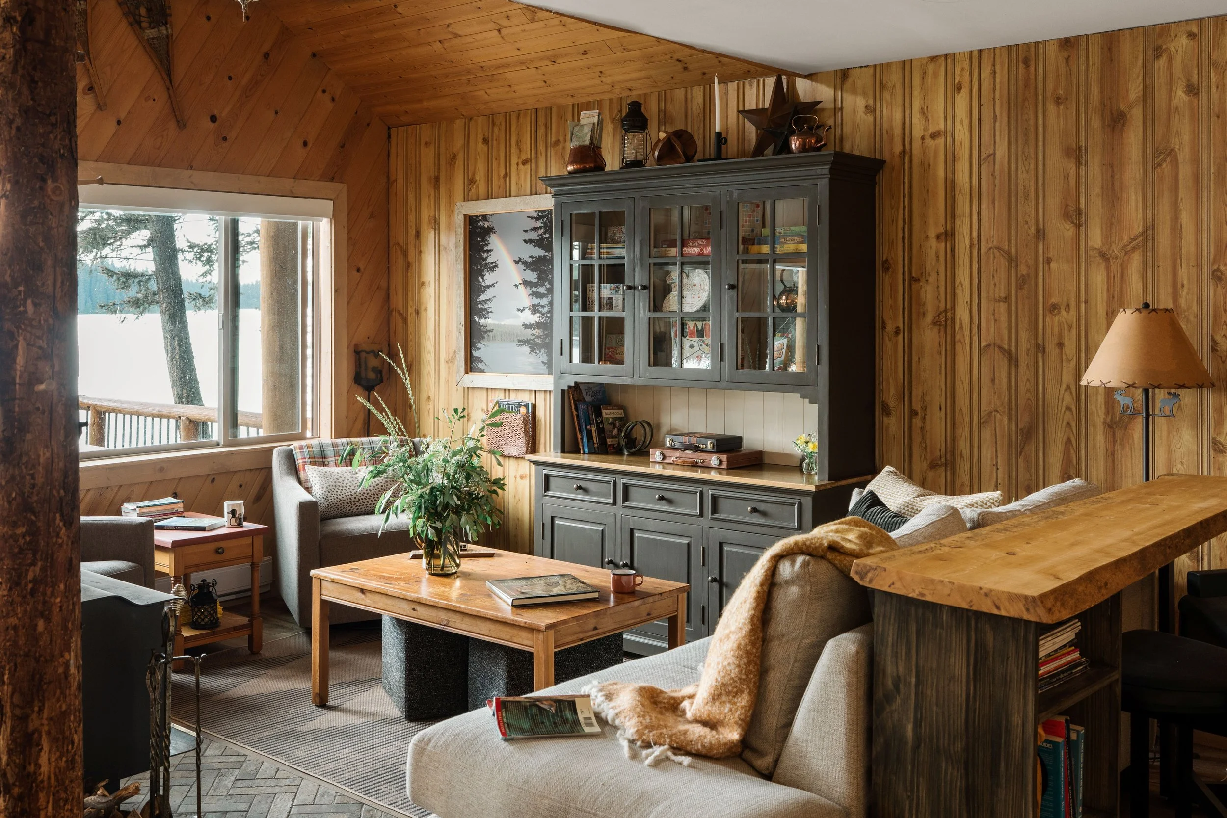 A dark grey sideboard and tall hutch is a focal point in a living room as it is host to all of the cabin's games. On top of it sits a collection of antiques and copper pots. Vintage backgammon boards sit on top of the wood top. Pine coffee table.