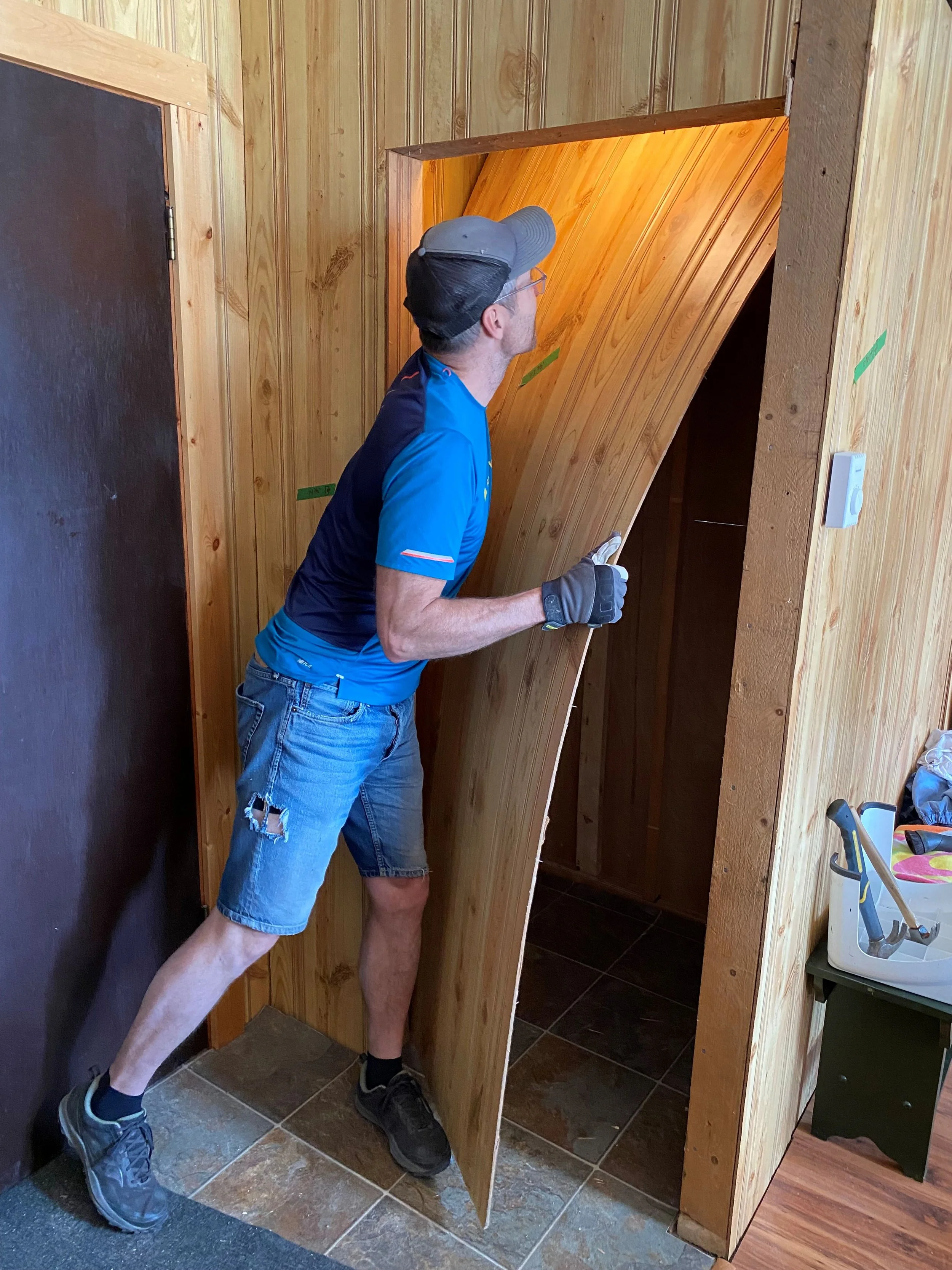 A tall man wearing work gloves is bending a faux wood 4x8 sheet of paneling. Removing it from a closet space. He is being careful to not damage the light above inside the closet and is using his foot to lean the bottom against, so it bends gently.
