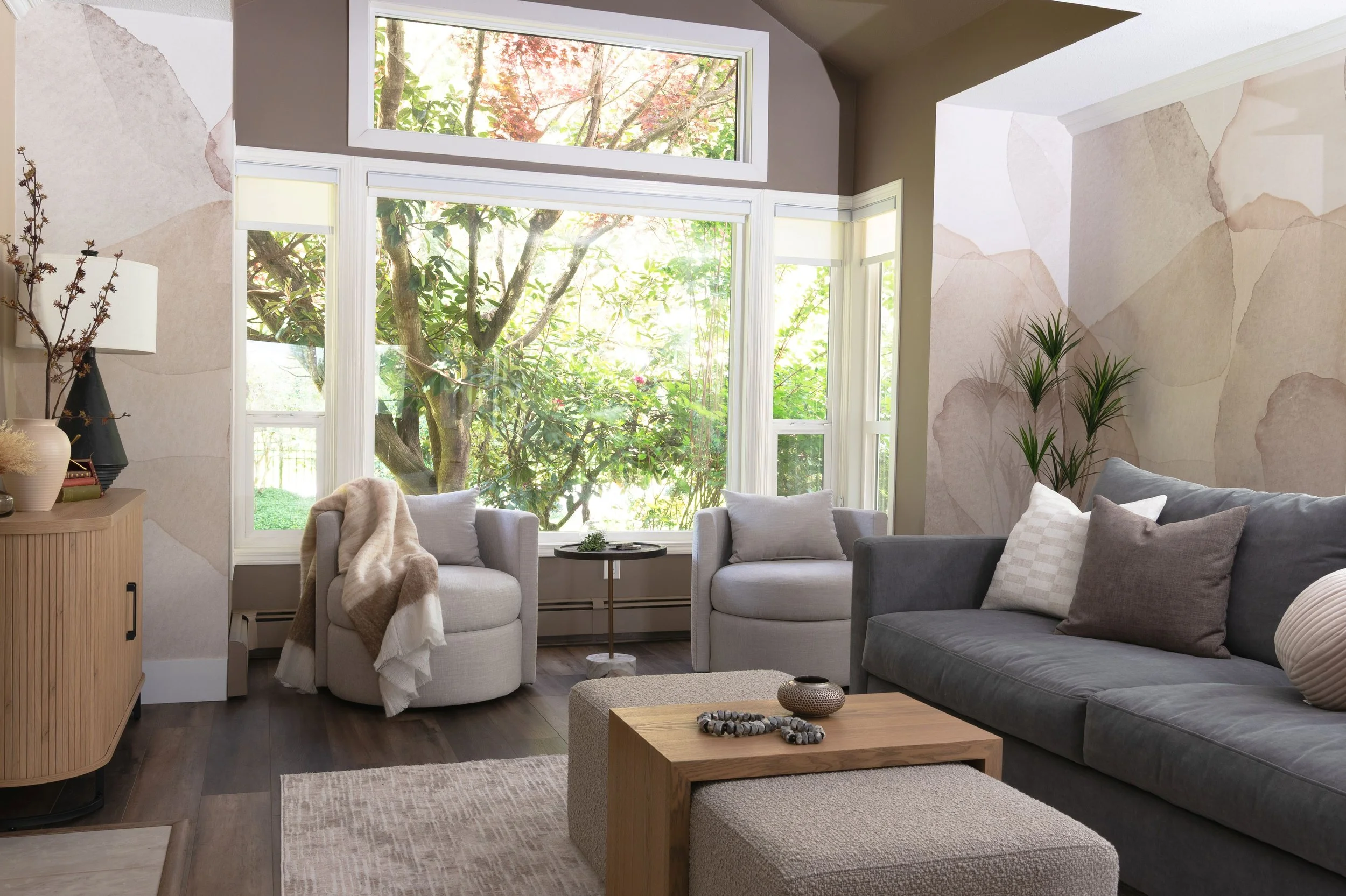 A calm, newly updated living room receives new bold brown Sycamore Tan paint color in the vaulted peaked ceiling in front of large bay windows. Two light grey swivel chairs and round side table are in front of the bay windows. White oak furnishings.