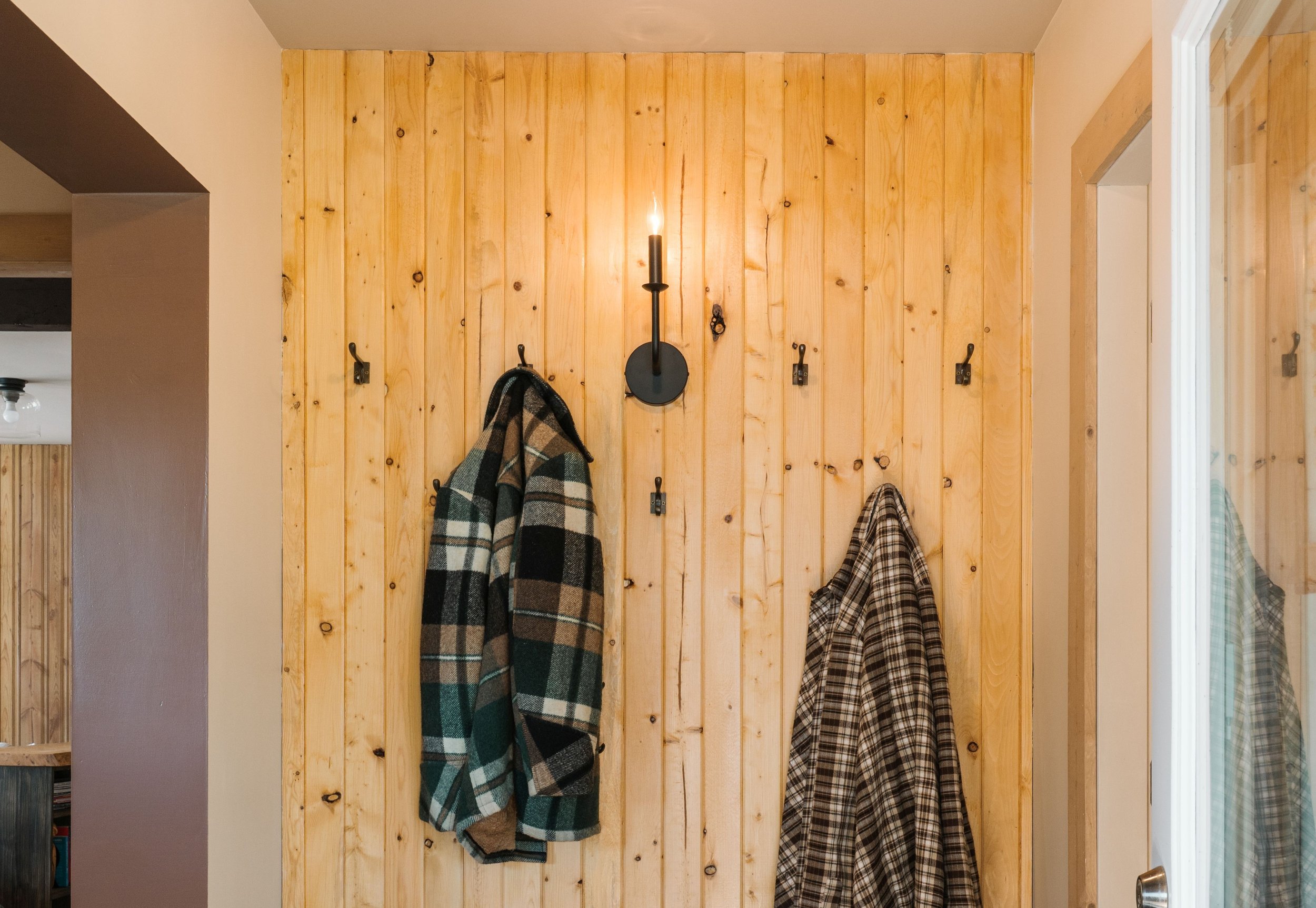 A cabin entrance greets visitors with a pine t&g tongue and groove feature wall with antiqued Etsy wall hooks. Also a single modern black candle wall sconce  is centred above the hooks. Plaid country jackets and coats hang from the hooks.