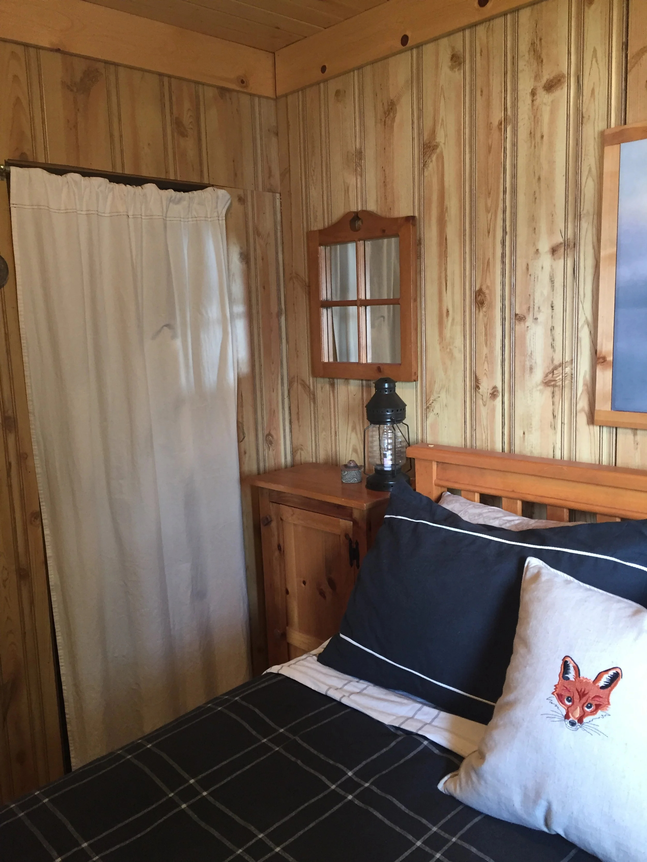 A white canvas fabric is used as a curtain for a closet door in a tiny cabin bedroom. An embroidered fox is on a toss cushion. The bedroom furnishings are pine.
