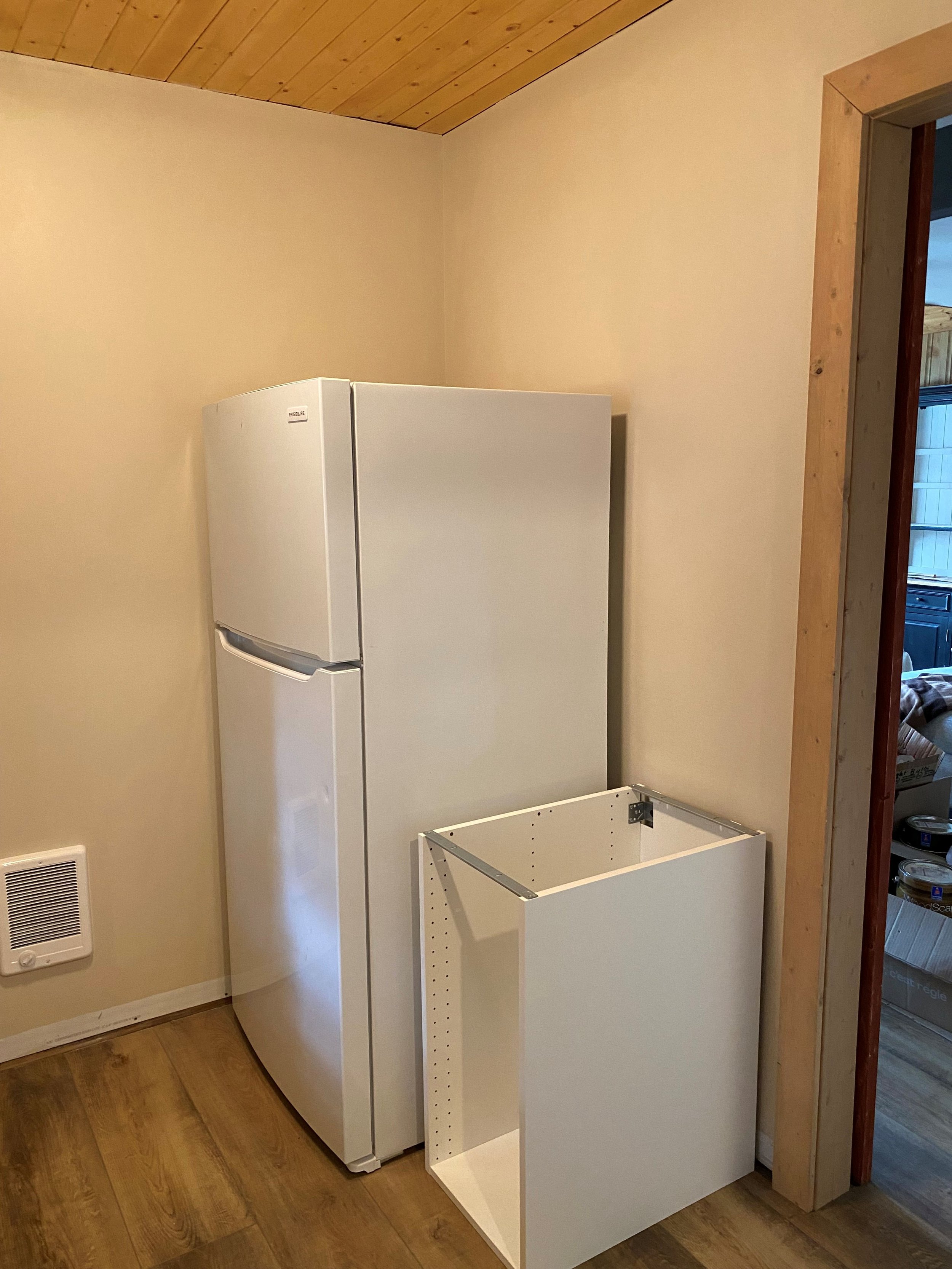 A white fridge with upper freezer is an extra fridge in a storage room in a cabin setting. A IKEA cabinet sits to the right of the fridge and a wall mounted heated is on an adjacent wall to help warm up this small room.