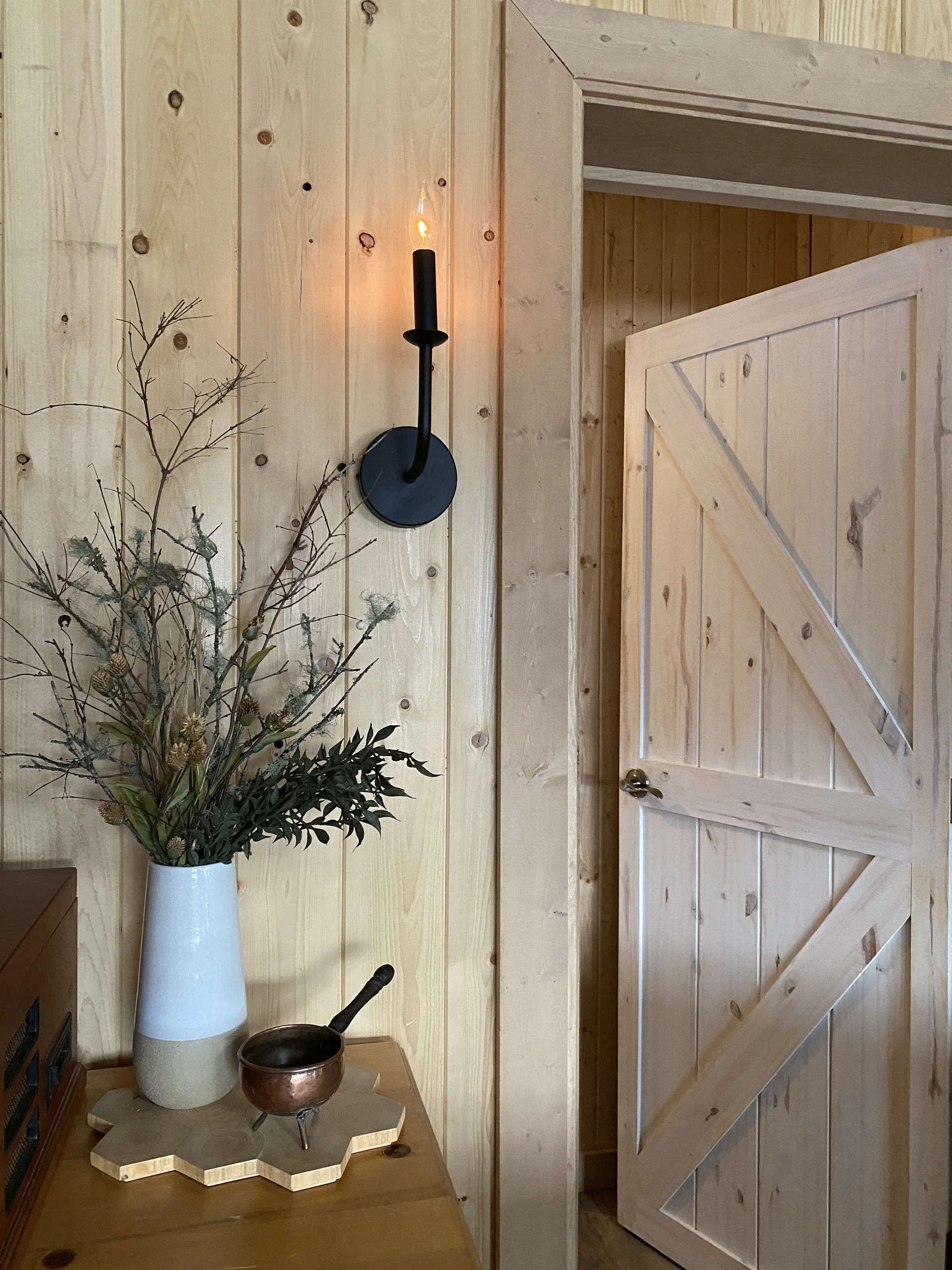 A modern, single black candle wall sconce contrasts against the rustic naughty pine wall. The warm light of the bulb illuminates the space. To the left is a vase of dried flowers and to the right is a doorway with a barn door into a bedroom.