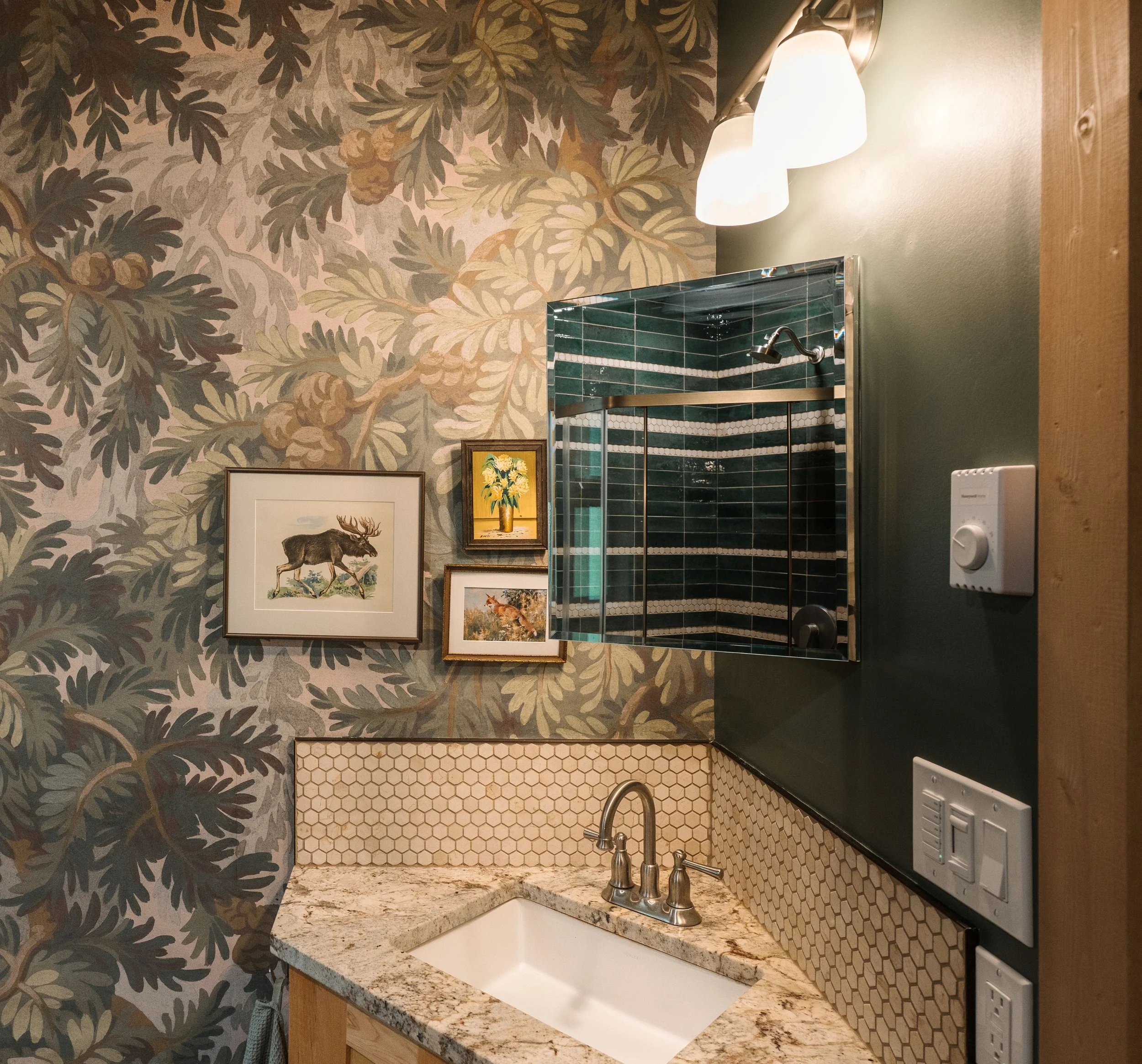 A cabin bathroom features one wall with Spruce Forest by Rebel Walls in green. Vintage art gallery wall with 3 brown frames hangs above a vanity that has small white hex as a backsplash. The recessed medicine cabinet miror shows the shower relection.