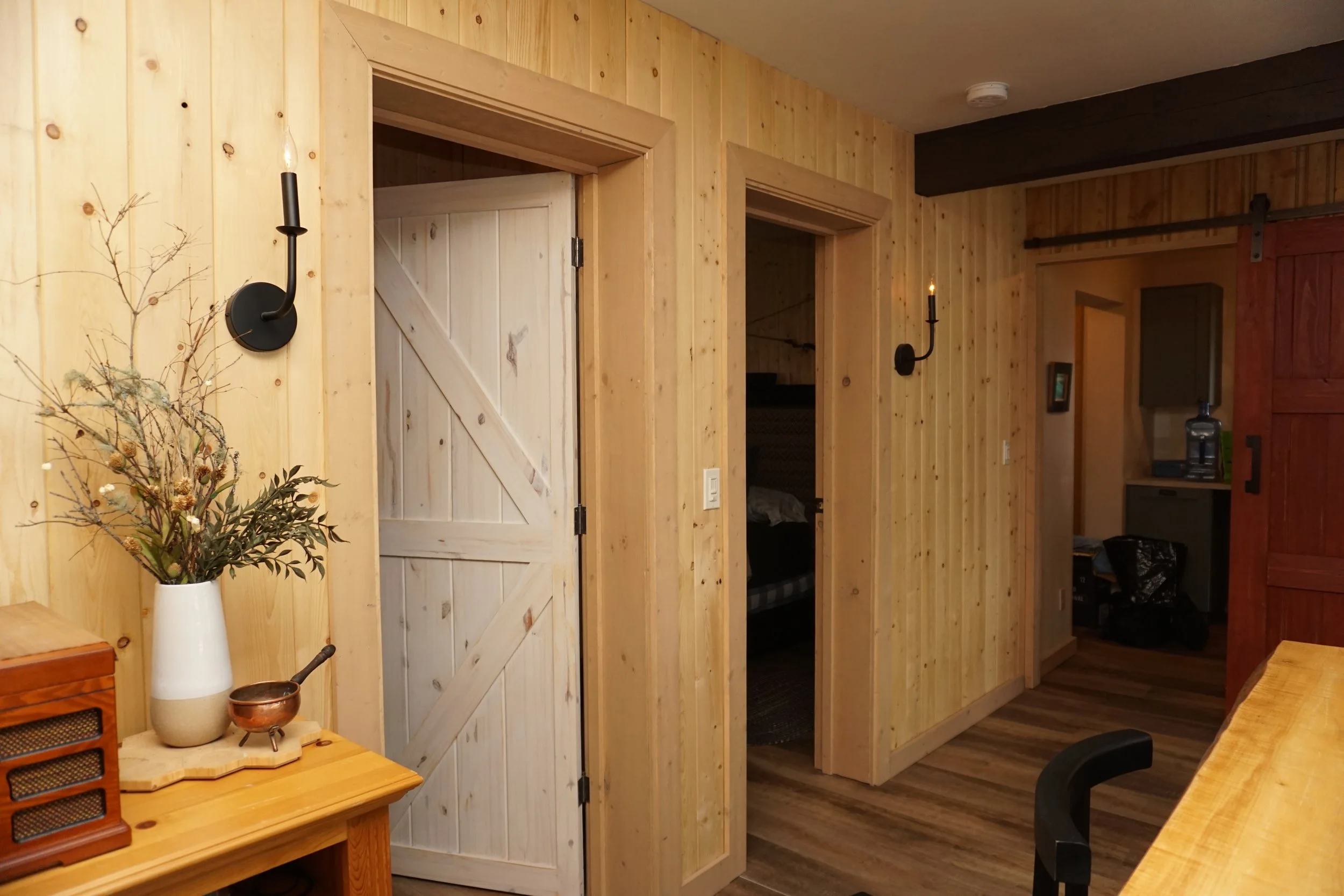 A hallway outside of two adjacent bedrooms has two black candle wall sconces on the wall, illuminating the walk to the room in the distance. It is a cabin setting with pine tongue and groove boards on the walls and the white-washed doors.