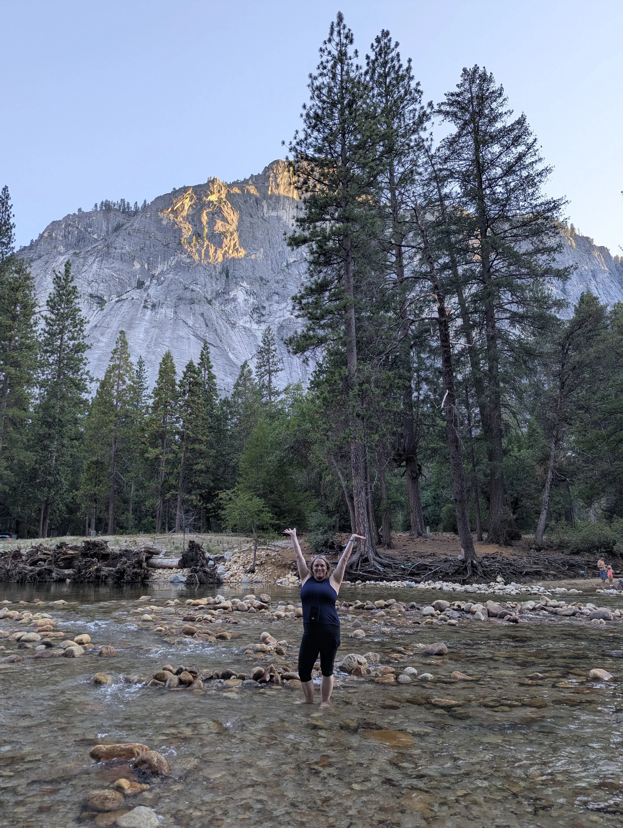 Strong woman stands under mountains in Yosemite National Park. She is barefoot and in a creek with her arms lifted overhead for joy.