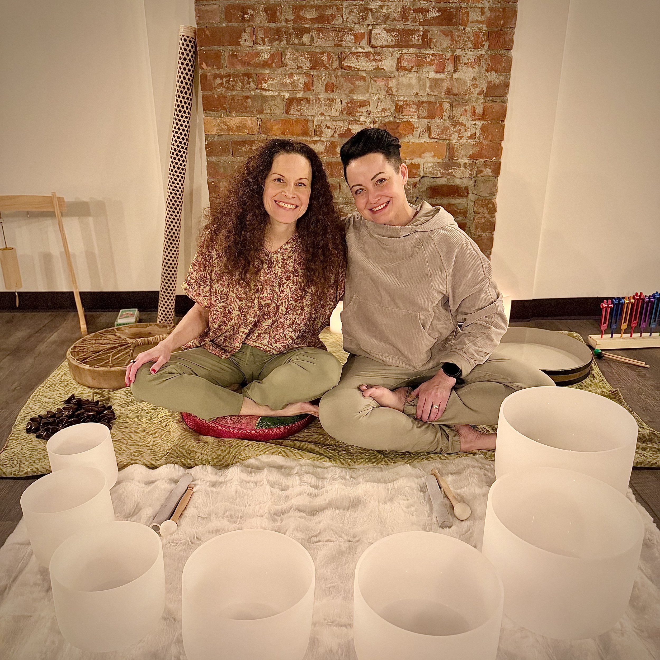 Two women sitting cross-legged on a plush surface, smiling at the camera. They are surrounded by singing bowls and other musical instruments, with a brick wall in the background.