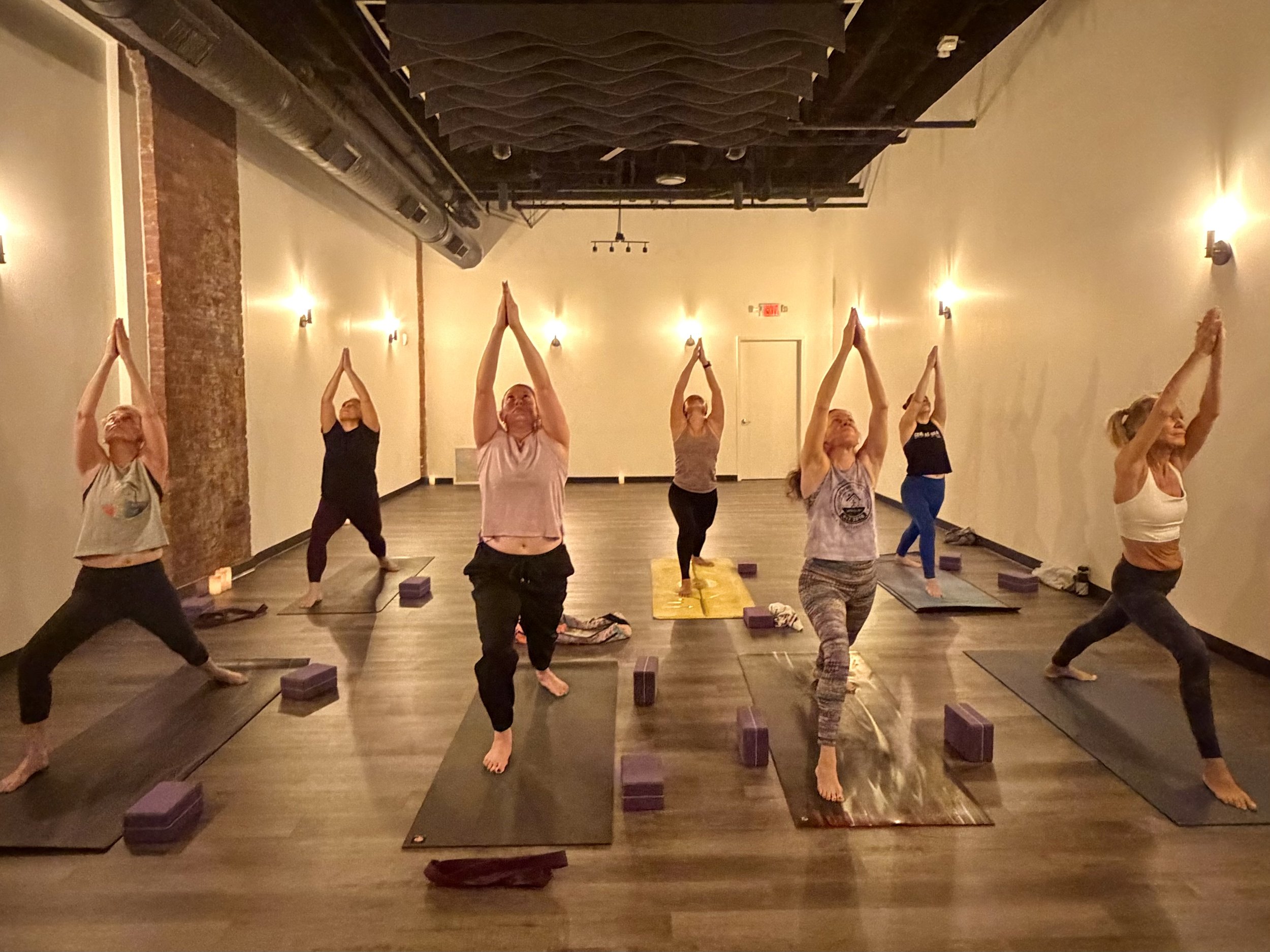 A group of people practicing yoga in a dimly lit studio with wooden floors and warm lighting, standing on yoga mats with their hands raised above their heads and palms together.
