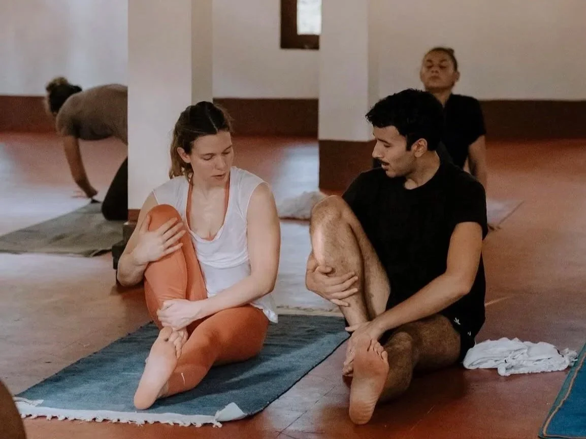 Two people sitting on yoga mats in a yoga class, holding their legs in a stretching pose, with two women in the background stretching.