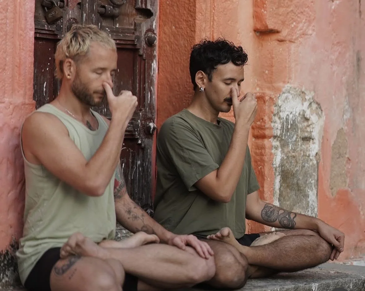 Two men sitting cross-legged on the ground, meditating with their eyes closed, against an orange wall.