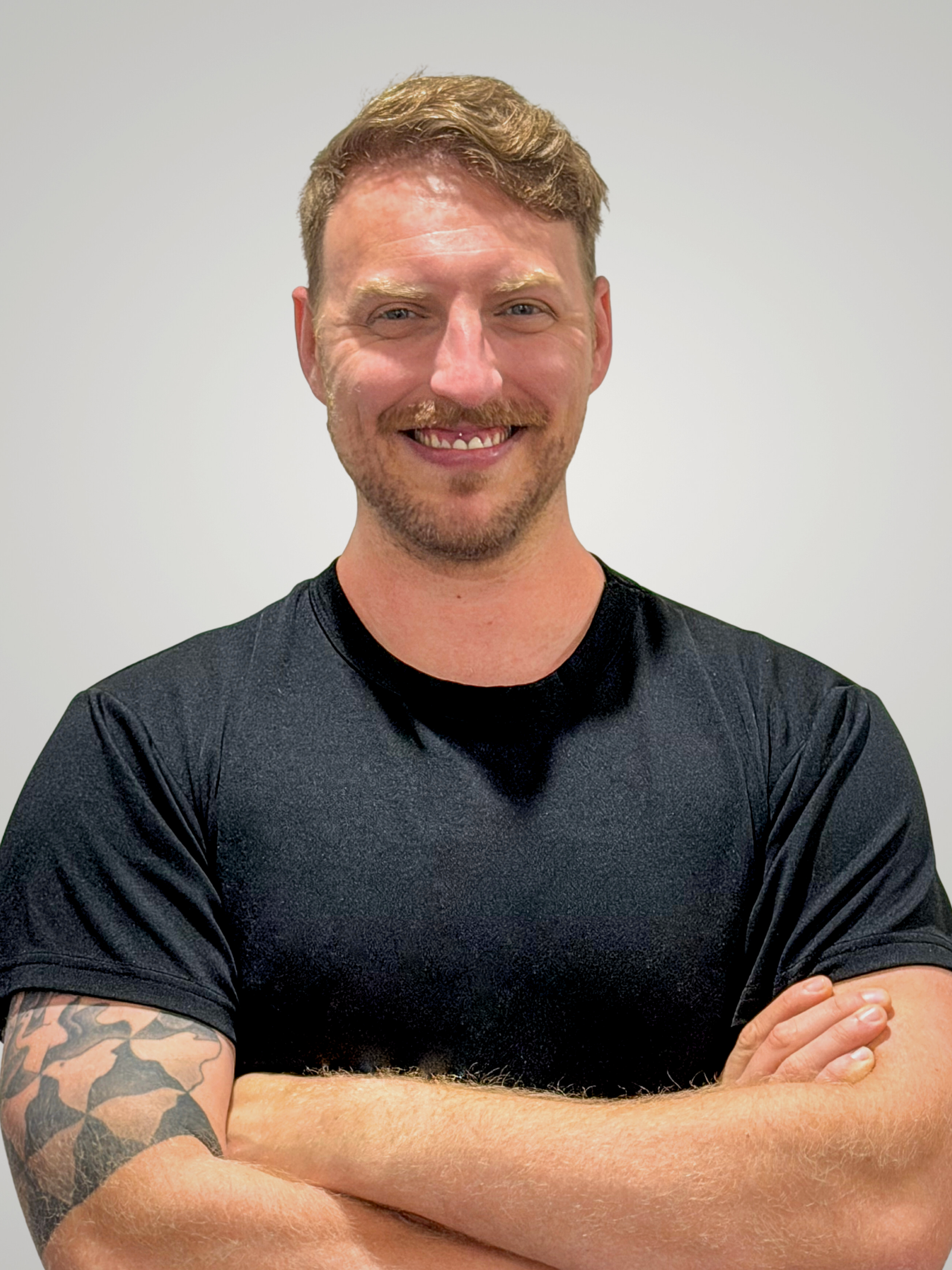 A smiling man with short, light brown hair and a beard, wearing a black athletic shirt, standing against a plain light-colored wall.