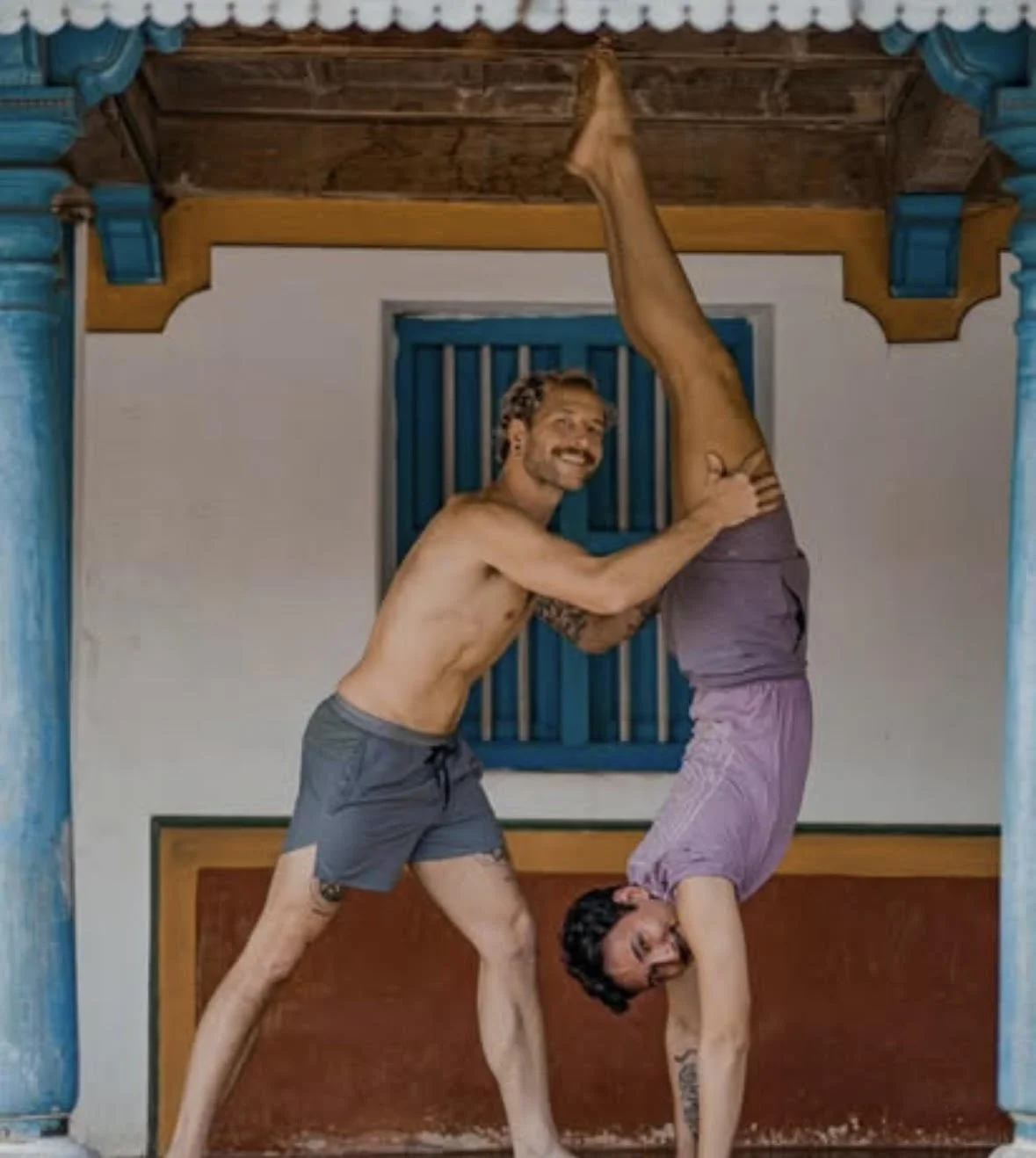 Two men practicing acro yoga indoors, with one man balancing upside down and the other supporting him, both smiling.