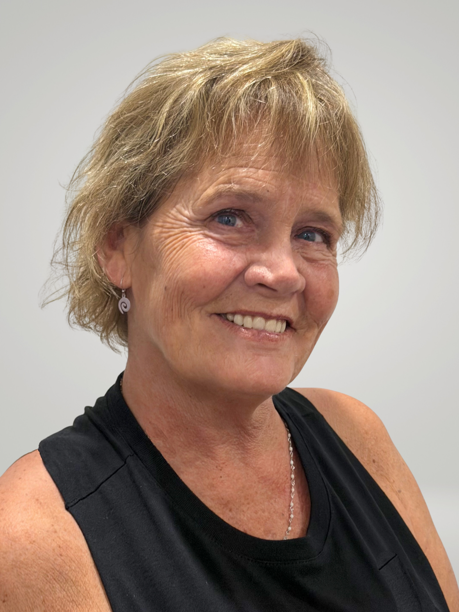Close-up of a smiling woman with short, curly blonde hair and blue eyes, wearing a black sleeveless top, earrings, and a necklace, against a plain light-colored background.