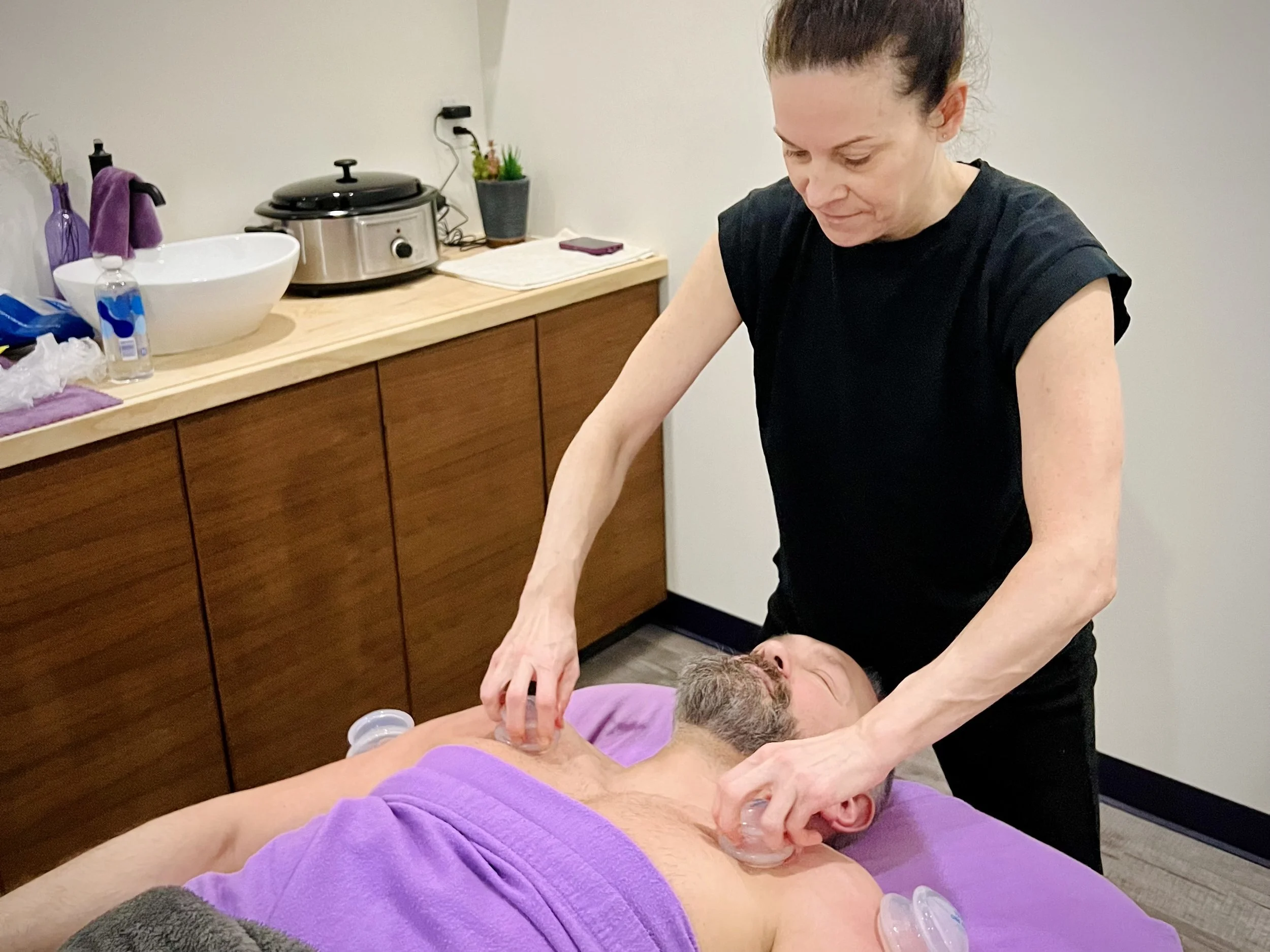 A woman giving a massage to a man lying on a massage table, with a purple towel covering his waist, in a room with a wooden counter and a sink in the background.
